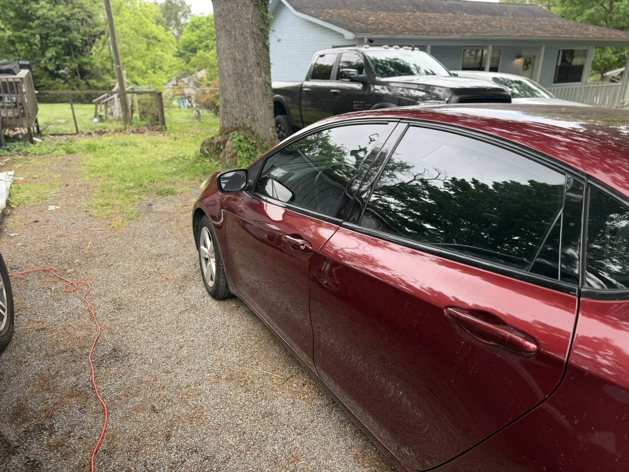 A red car is parked on a gravel driveway, with a black pickup truck behind it and a house with a porch in the background. A tree with a vine around its trunk is visible, and a red extension cord is on the ground.