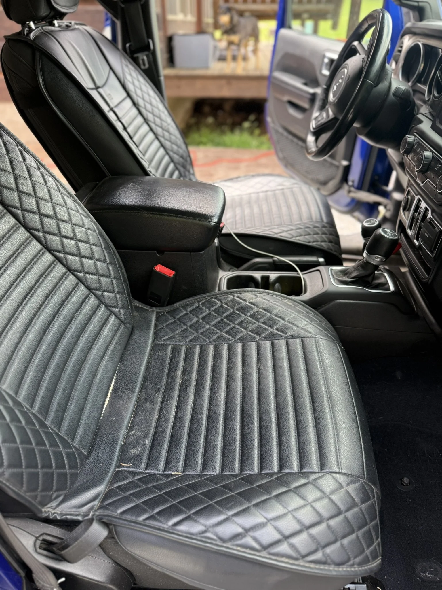 Inside a vehicle, the black leather seats with a quilted diamond pattern, center console, steering wheel, and parts of the dashboard are visible, with a blurred background showing an outdoor setting.