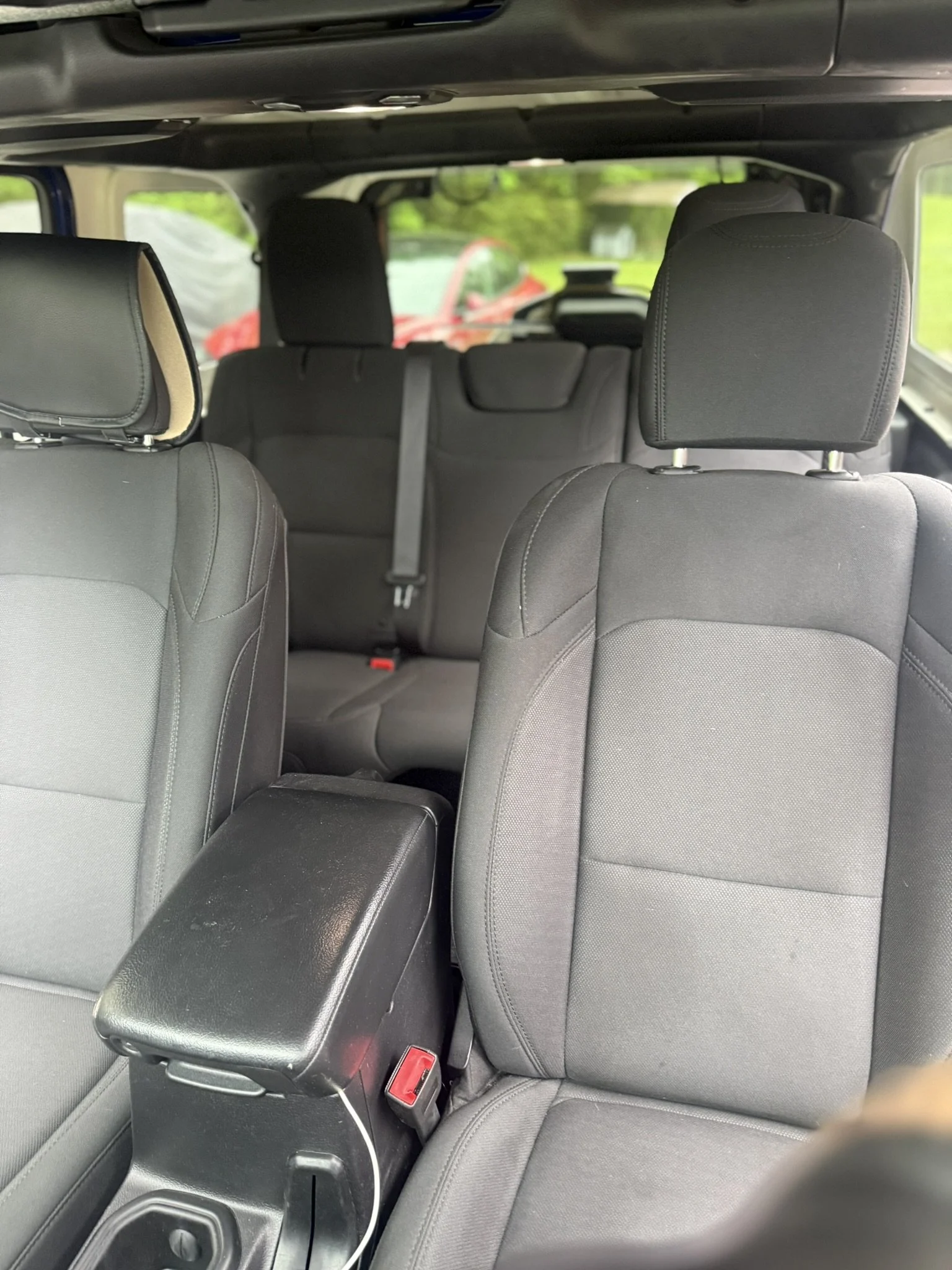 Interior of a vehicle showing the back of front seats and the rear seats with headrests. A black armrest with a red latch is visible between the seats, and a device with a cable is attached to it. The background shows outside greenery and a red car.