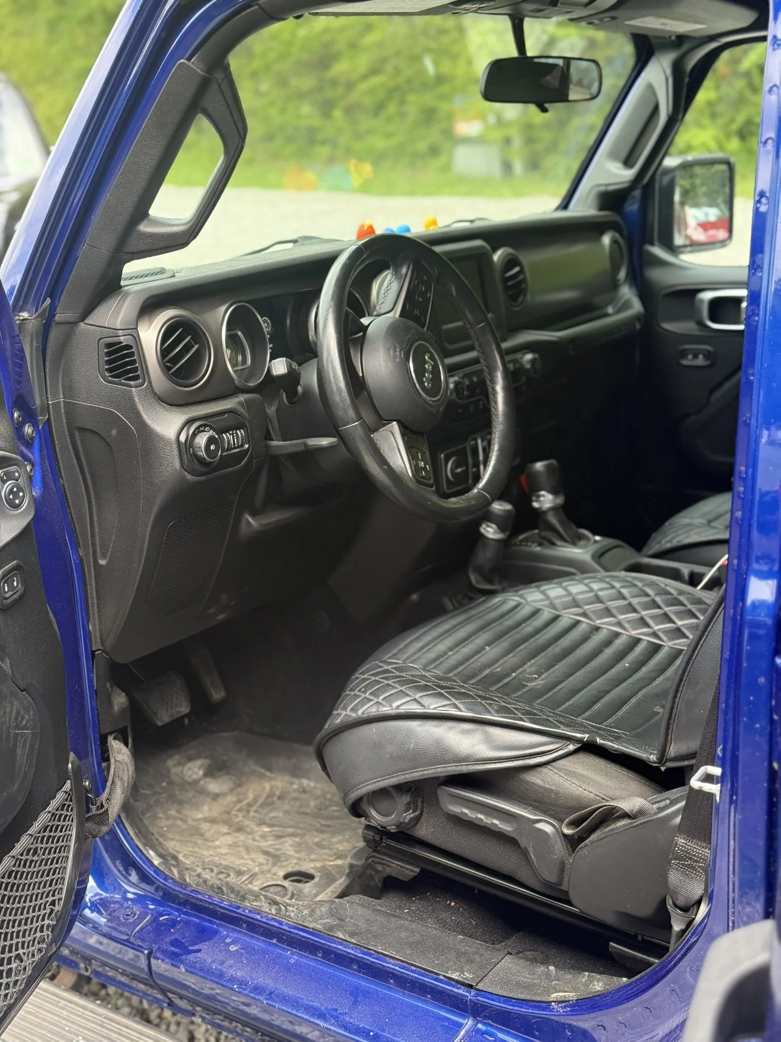 Interior of a blue Jeep vehicle, showing the driver's seat, dashboard, steering wheel, gear shifter, and part of the passenger seat and door, with a green outdoor background visible through the window.