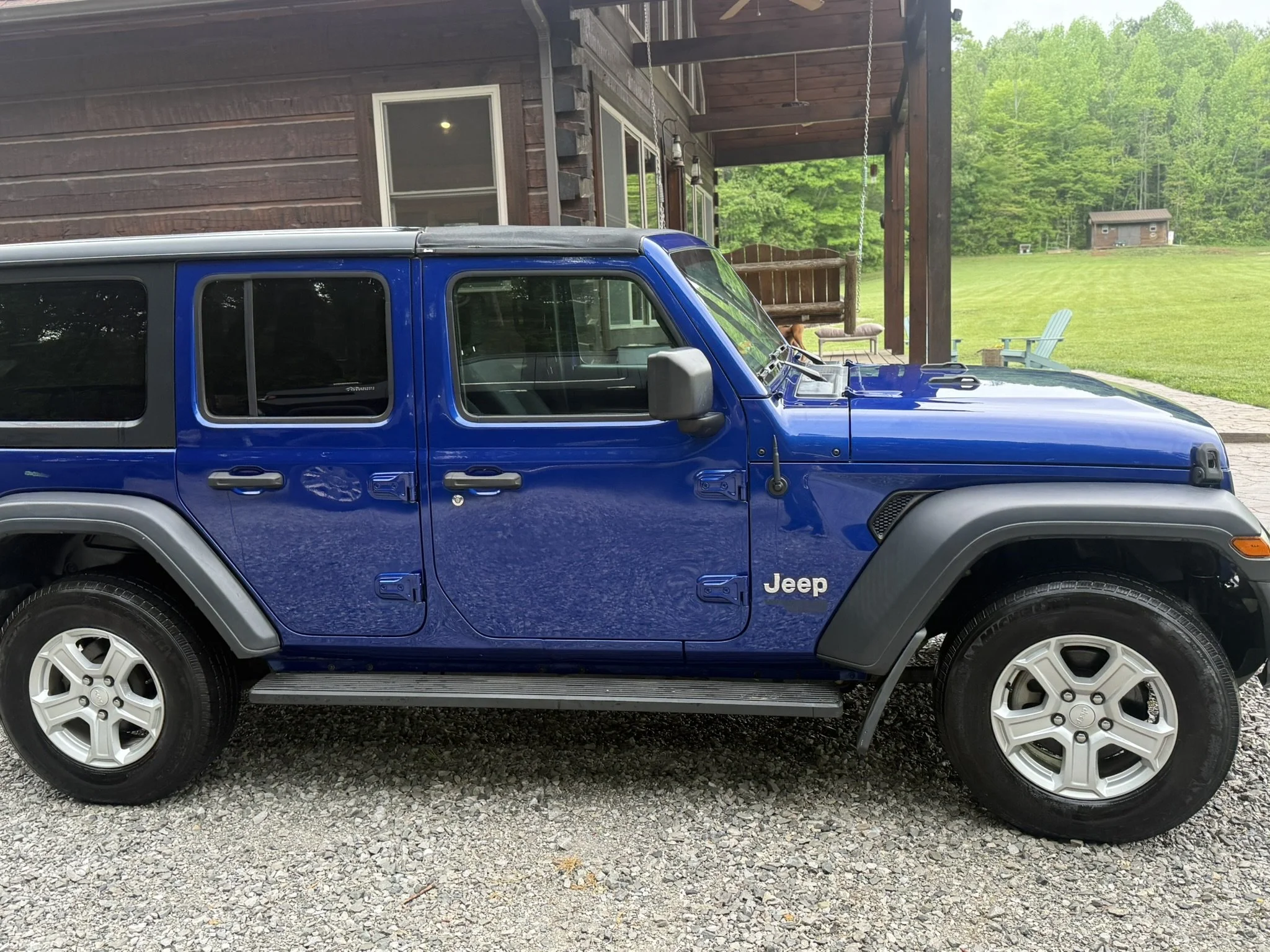 A blue Jeep parked on a gravel driveway in front of a house with a porch and swing, surrounded by green trees and open yard.