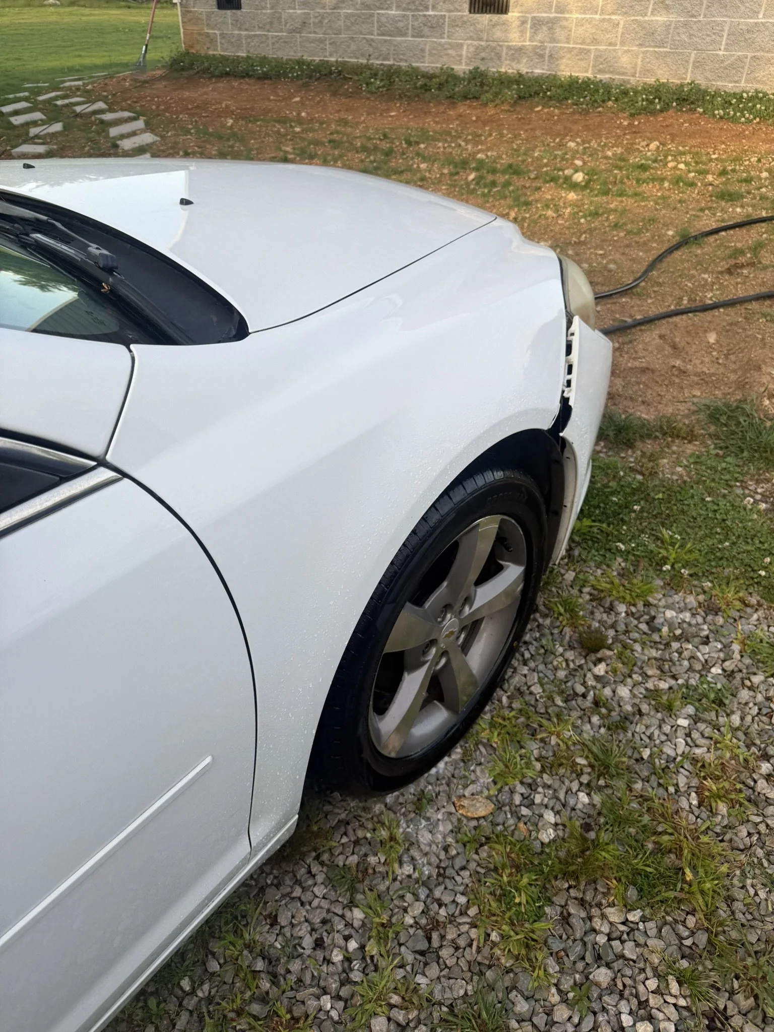 A white car is parked on a patch of grass and gravel next to a building. The front right side of the car is visible, with some damage to the bumper and the wheel. An electrical cord is plugged into the car's front bumper, presumably for charging.