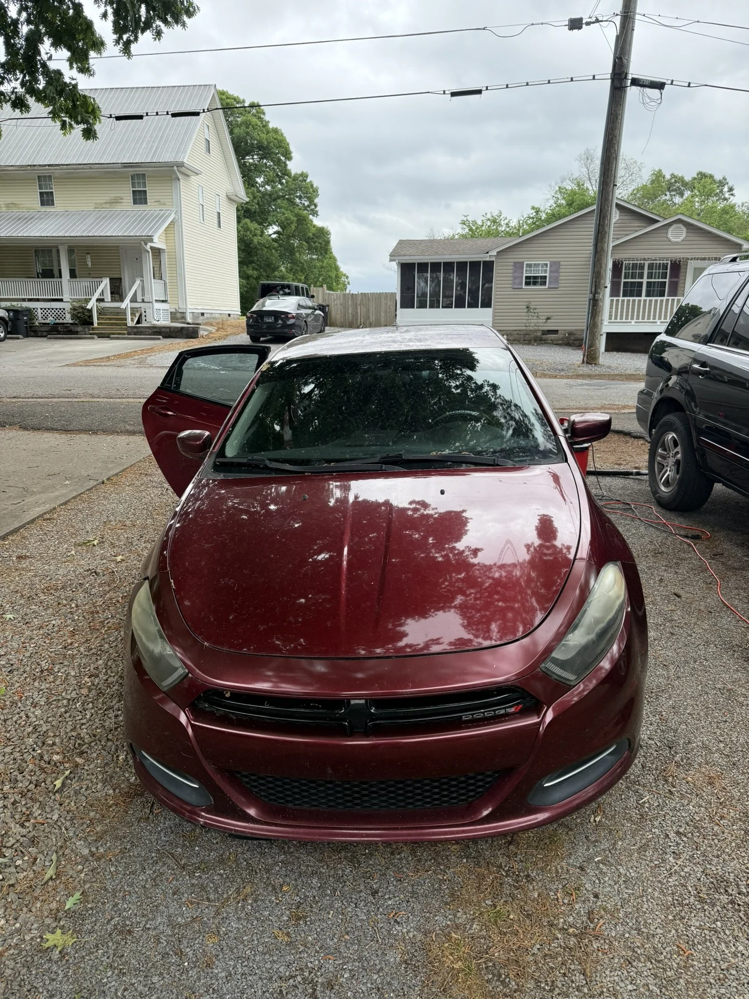 A red Dodge sports car parked on a gravel driveway with one door open, in front of suburban houses on a cloudy day.