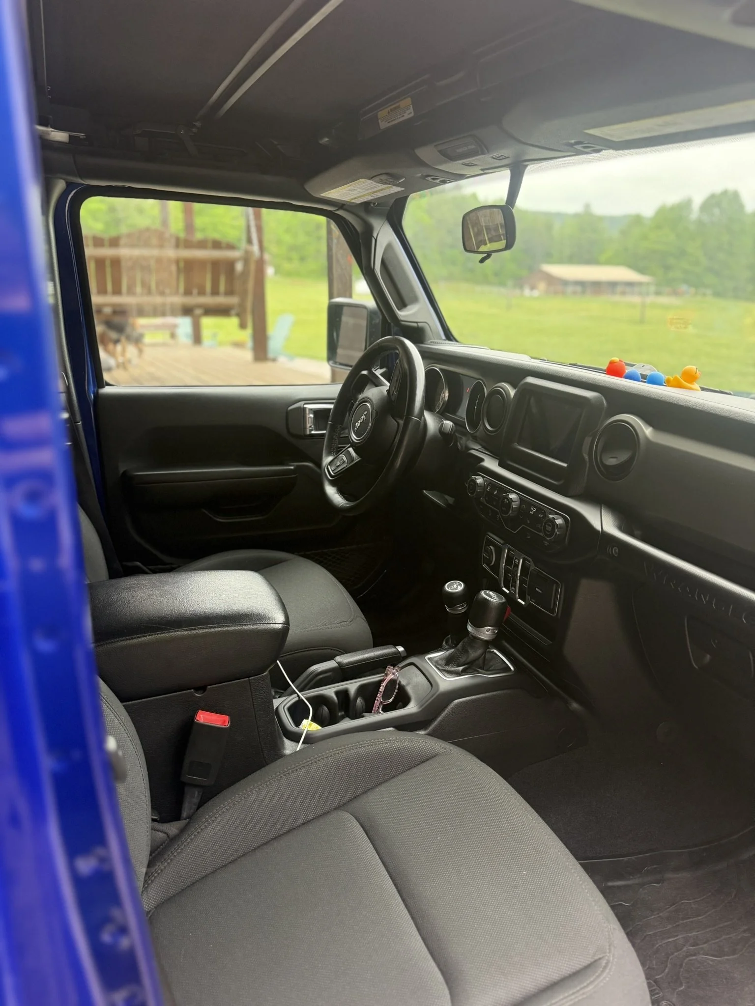 Inside view of a vehicle's front interior, showing the dashboard, steering wheel, gear shift, and front passenger seat with a window overlooking green trees and a house in the background.