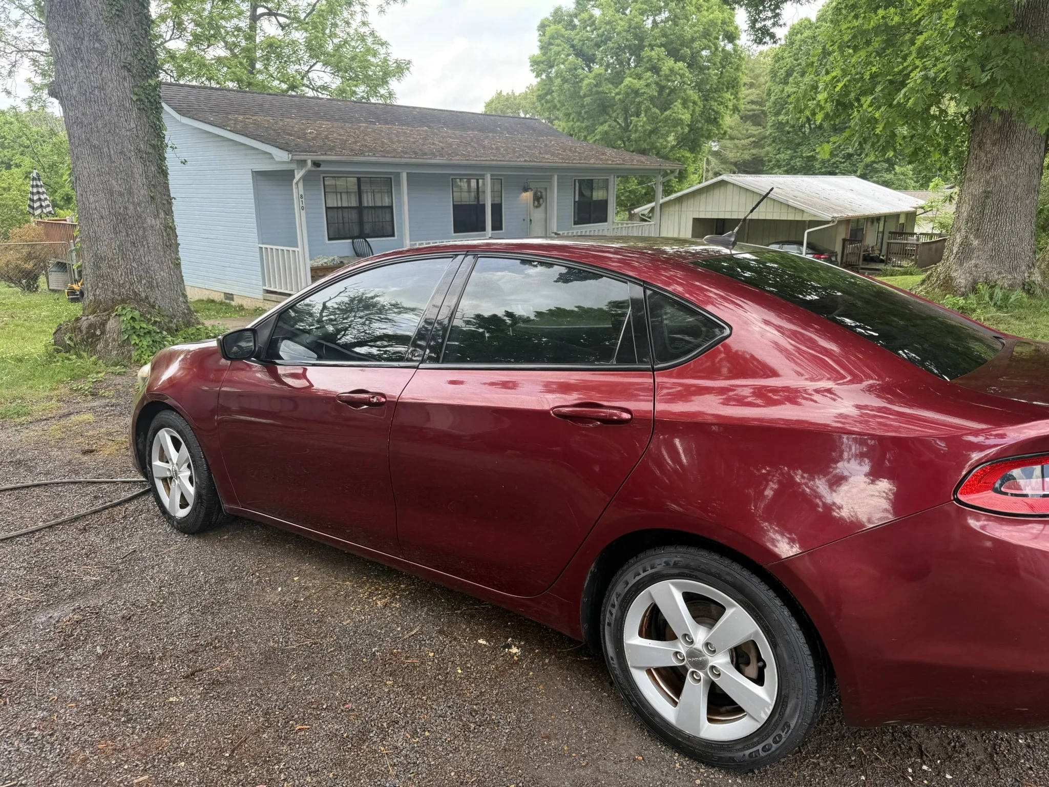 A red car is parked near a blue house with trees and another small structure in the background.
