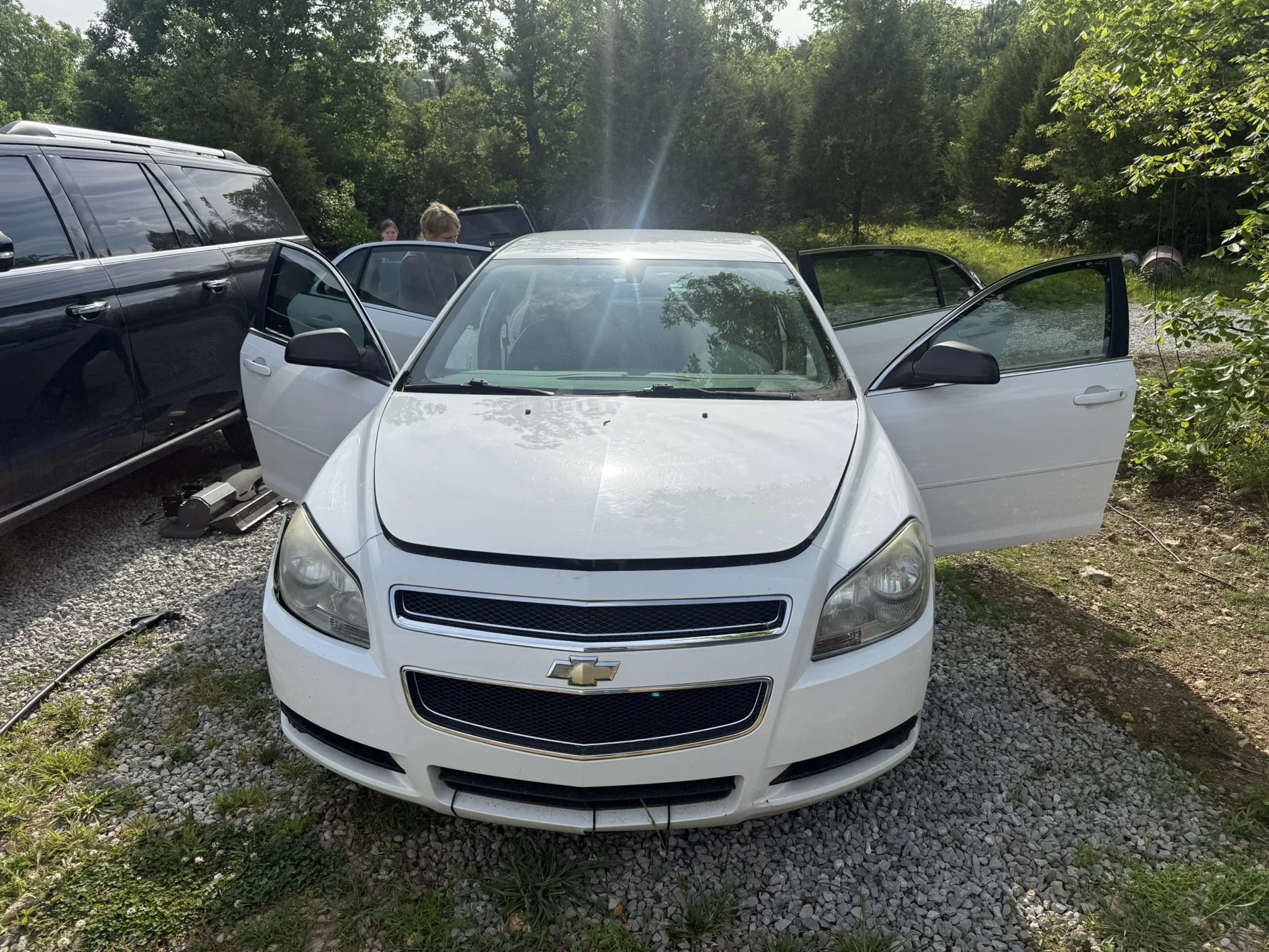 A white Chevrolet car with open front and rear passenger doors is parked outdoors among other vehicles in a wooded area with green trees, and some people are standing nearby.