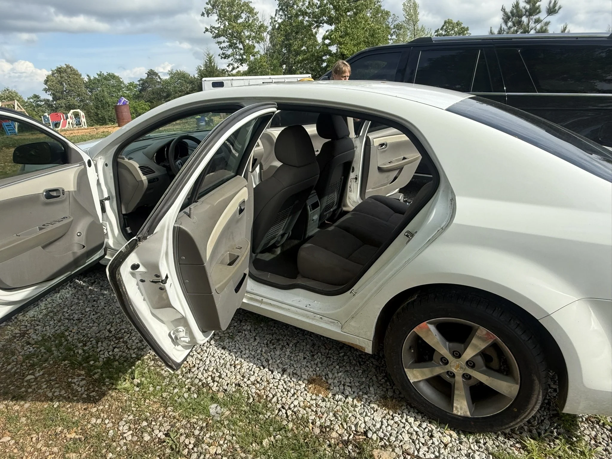 Interior of a white car with the front and rear passenger doors open, parked on a gravel area. The car's interior is beige and black, with black seats and beige panels. In the background, a man with light brown hair and a black shirt is visible, stan