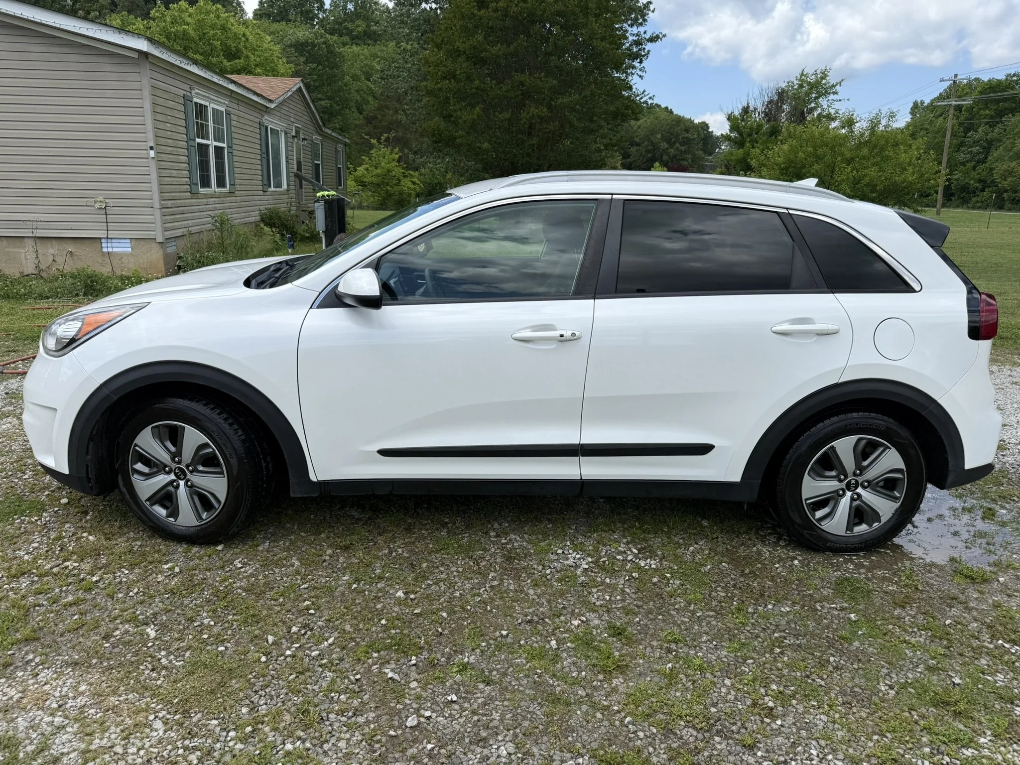 A white compact SUV parked on a gravel driveway with a house and green trees in the background.