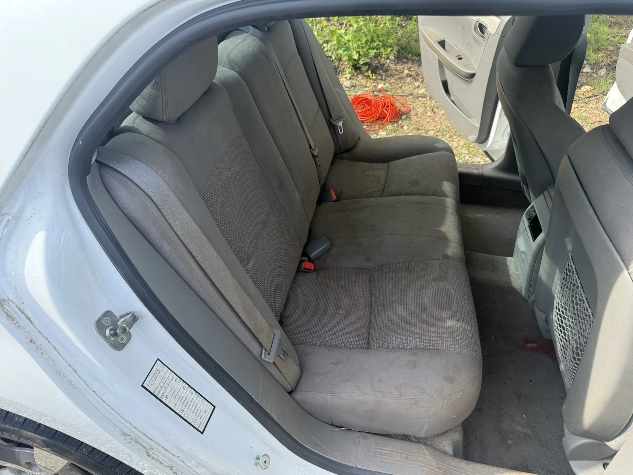 The back seats of a car with gray fabric upholstery. Outside the car, there is a pile of orange extension cords on the ground near some green bushes.