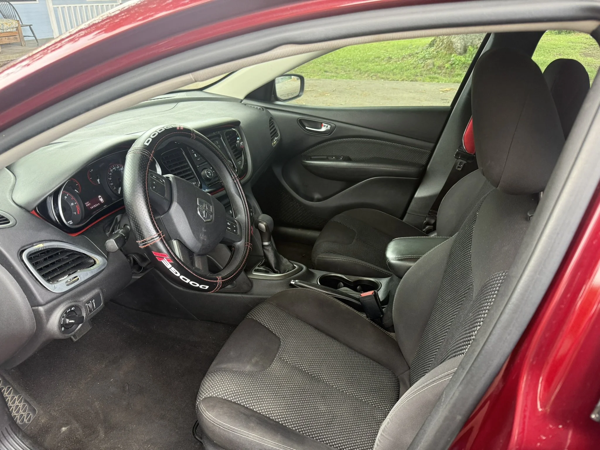The interior of a red car showing the driver's seat, passenger seat, dashboard, steering wheel with a Dodge logo, and gear shift. The seats are black with fabric and the dashboard is gray with various controls.