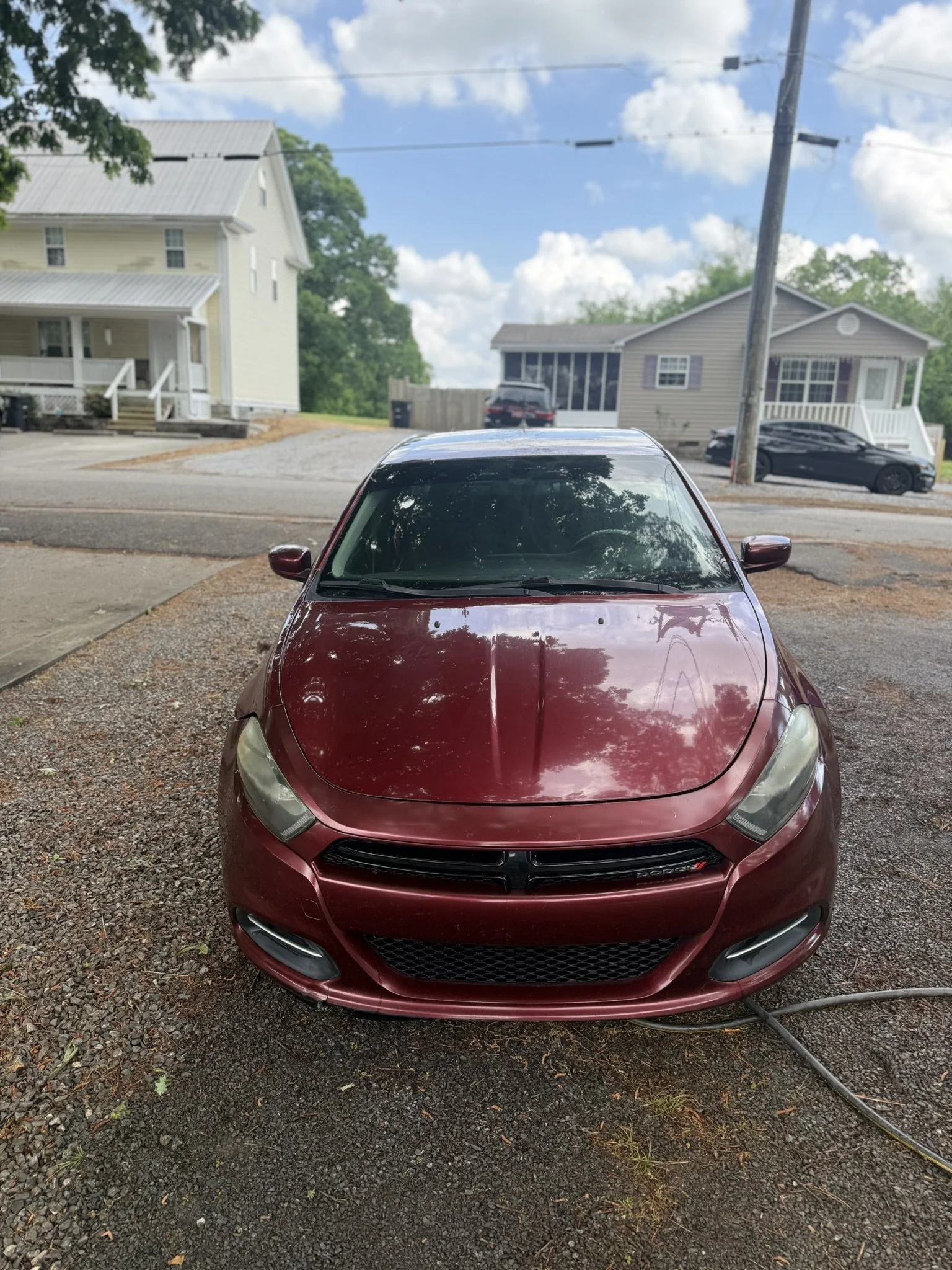 A red Dodge car parked on a gravel driveway in front of two houses, with trees and a blue sky with clouds in the background.