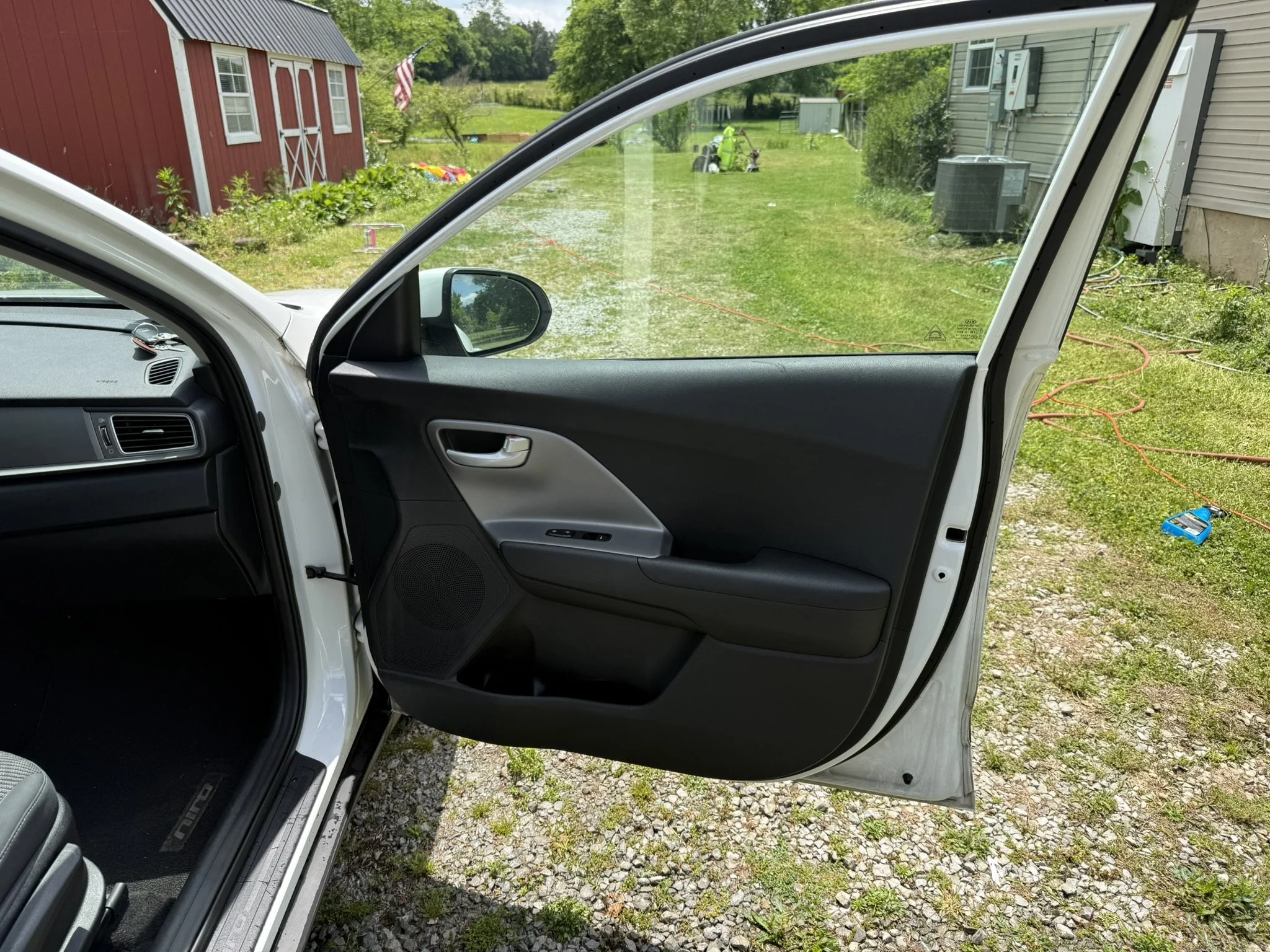 View from inside a car with the passenger door open, showing a grassy backyard with a red shed, a small yellow lawnmower, and a worker in bright yellow clothing trimming bushes. There are trees, an American flag, and some yard tools on the ground.