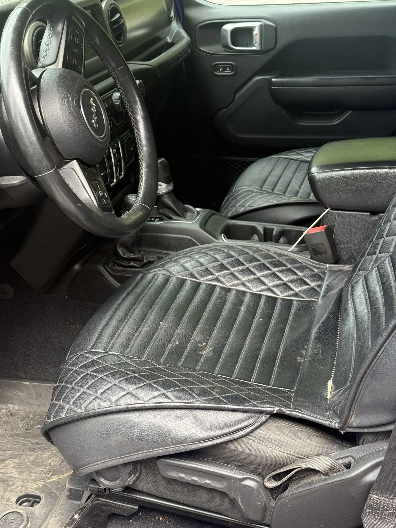 The interior of a vehicle shows a black quilted leather seat cover on the driver's seat, a Jeep steering wheel, and various dashboard controls. The door panel features a door handle and window controls.