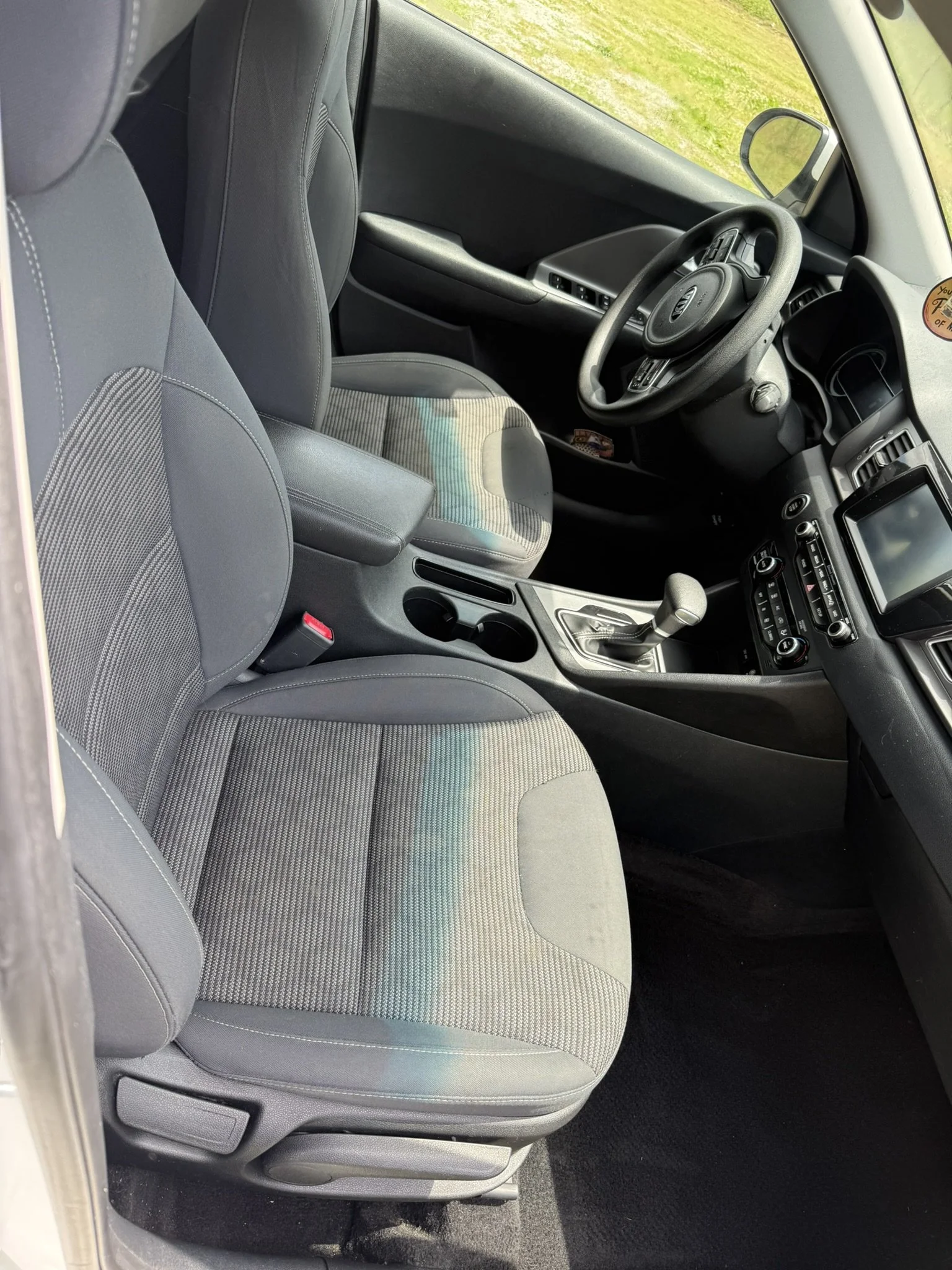 Interior of a car showing the front passenger seat, dashboard, steering wheel, and part of the driver seat, with some greenery visible through the windows.