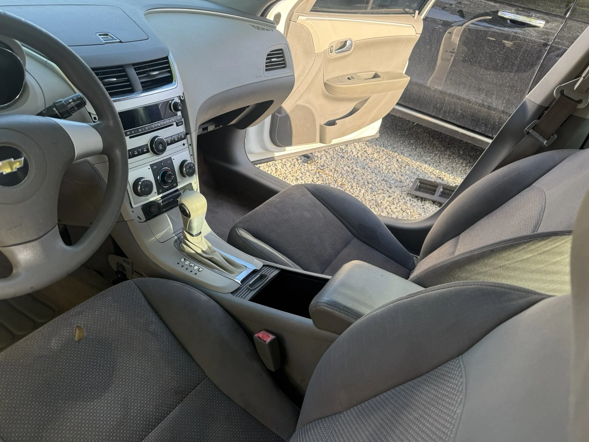 Interior of a vehicle, showing front seats, dashboard, and steering wheel, with visible window and exterior gravel ground.
