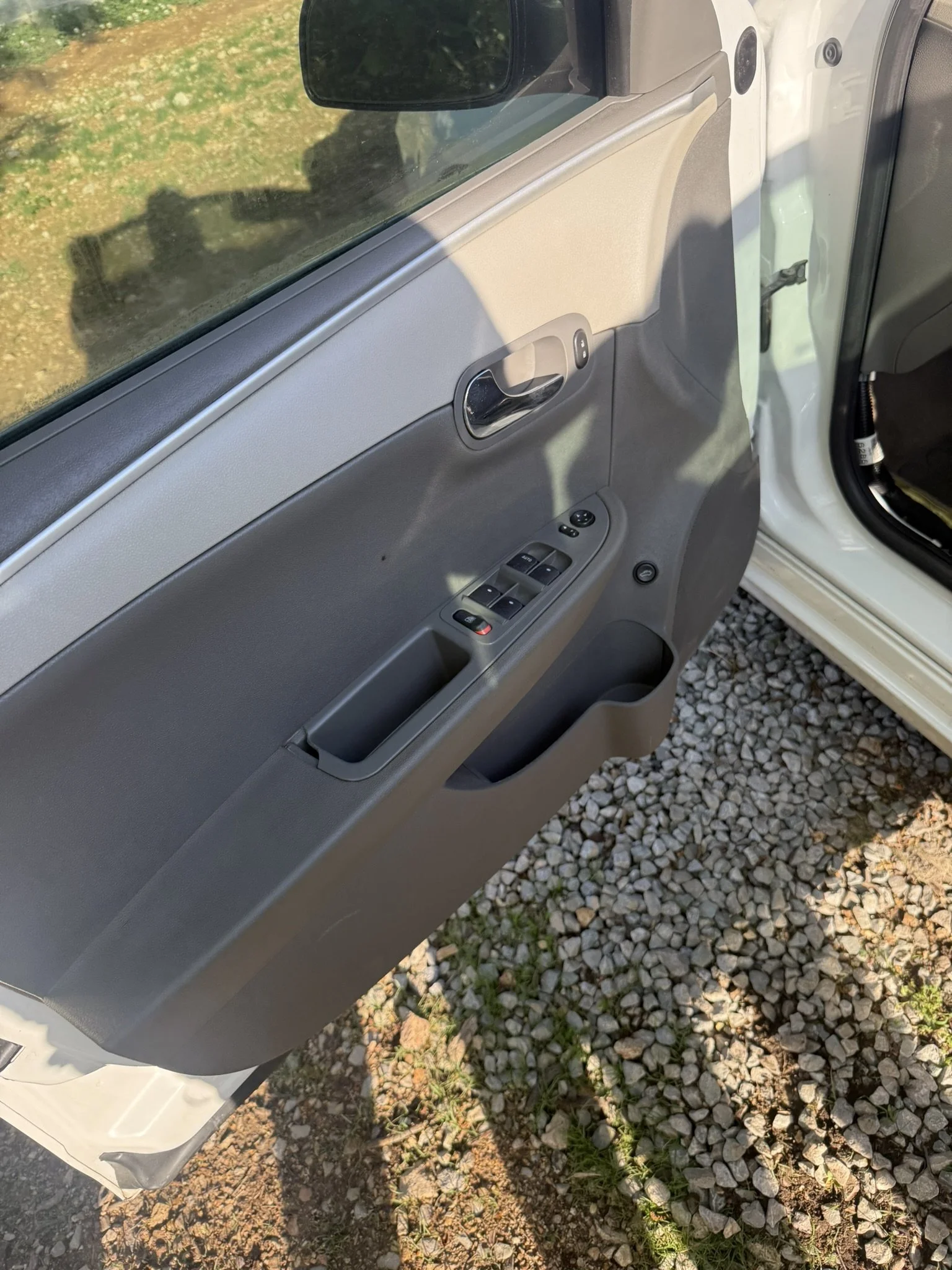 The interior door panel of a vehicle, showing power window controls, a door handle, and a window, with the vehicle parked on a gravel surface outside.