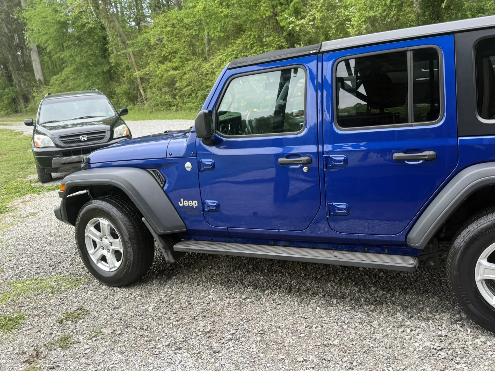 A blue Jeep parked on a gravel surface next to a black Honda SUV, with green trees and foliage in the background.