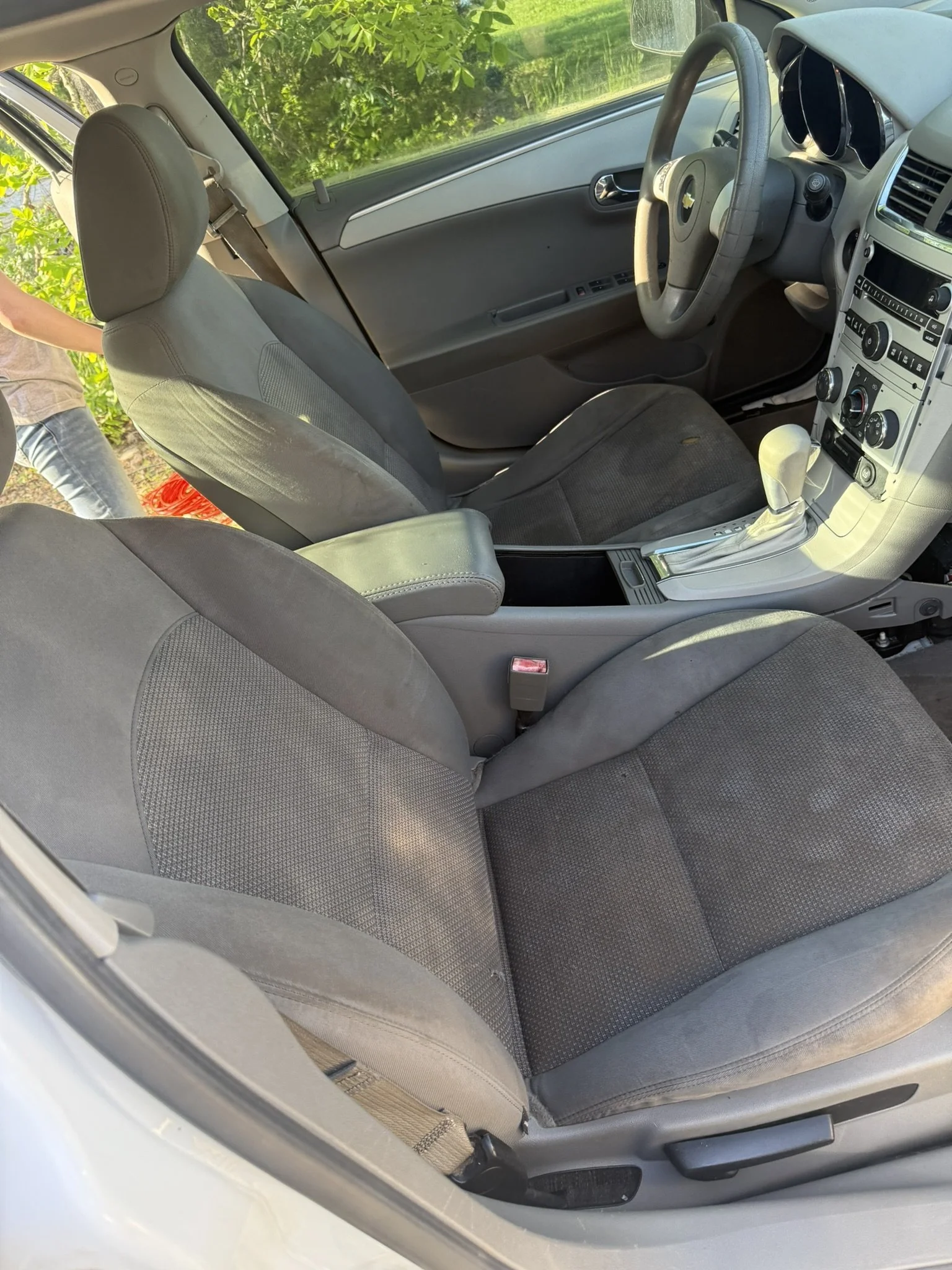 The interior of a car showing the driver's seat, passenger seat, dashboard, steering wheel, and gear shift, with greenery outside the windows.