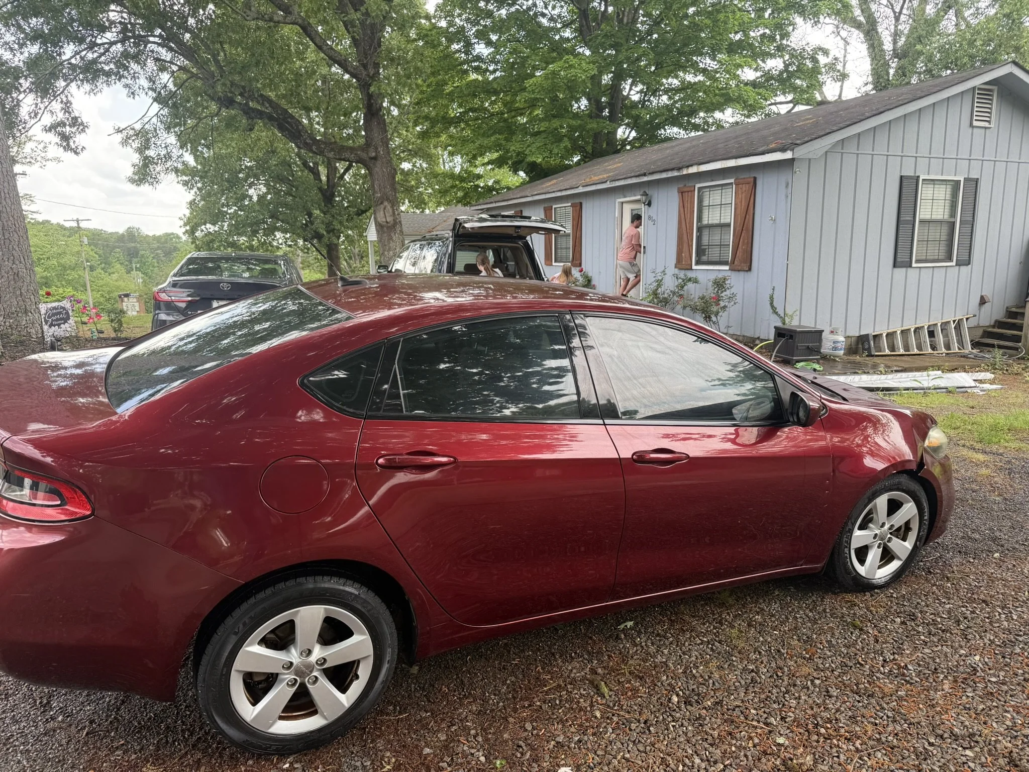 A red car parked in front of a gray house with brown shutters, with multiple other cars parked nearby under large green trees.