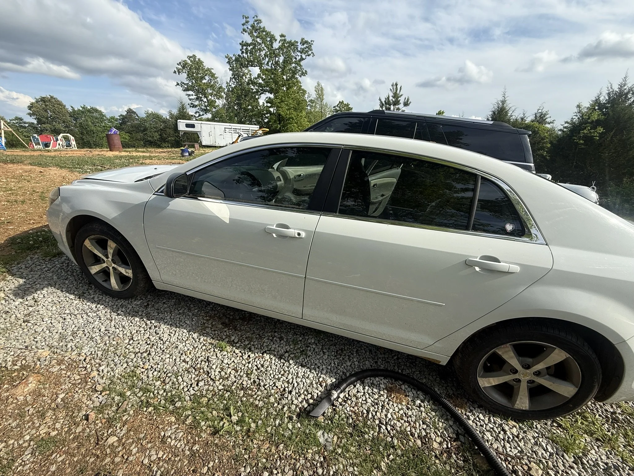 A white car is parked outdoors on gravel next to a garden hose. Behind it, a black SUV is visible, with trees and a cloudy sky overhead. In the background, there are children's play structures, a barrel, and a trailer.