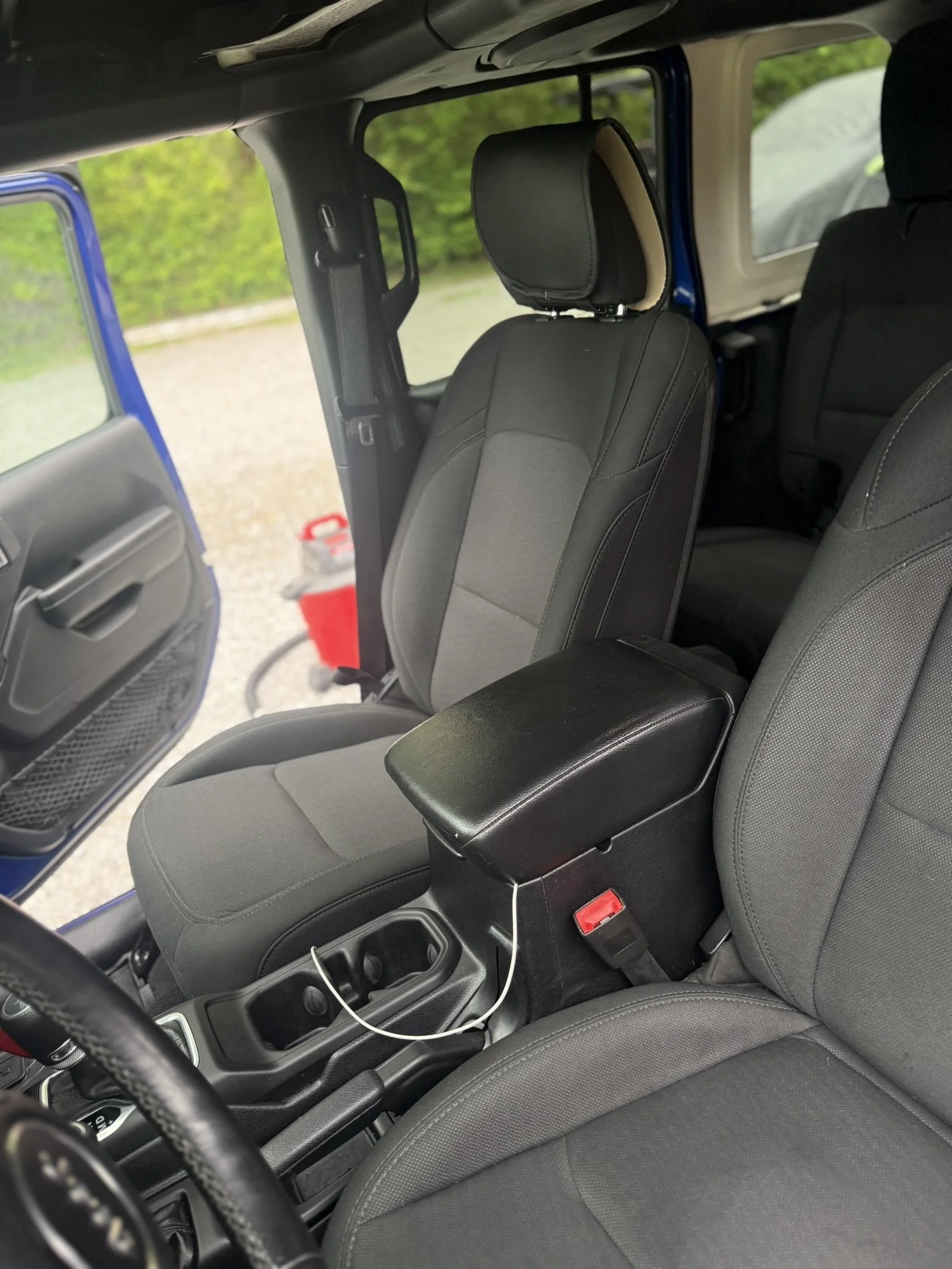 Inside a vehicle with the front seats visible, black interior, a charging cable plugged into a power outlet between the seats, and a red container outside the open door.