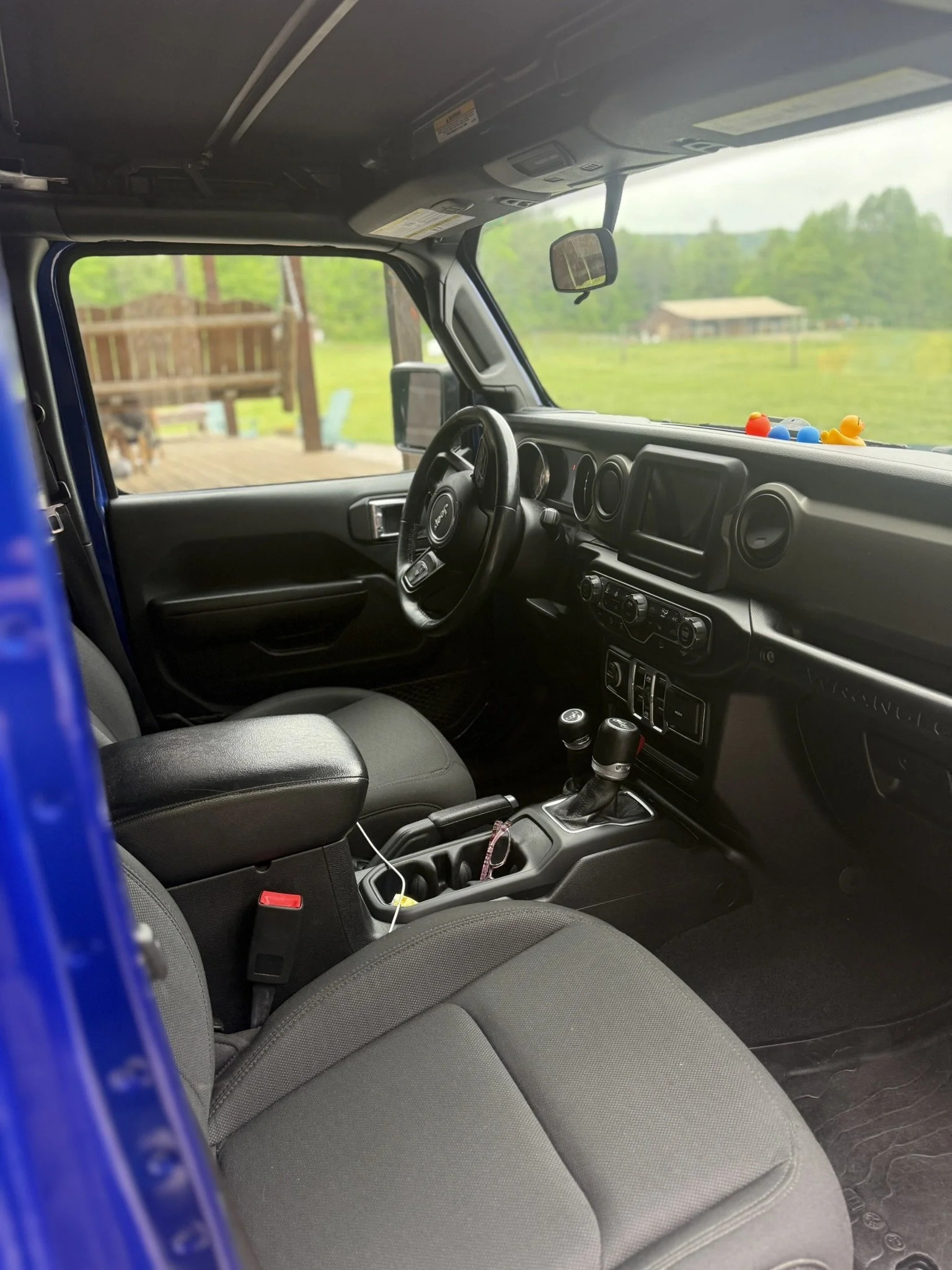 Inside a vehicle, showing the front passenger area with a dashboard, steering wheel, gear shift, and gray fabric seats, with a blurred outdoor scene through the windshield.