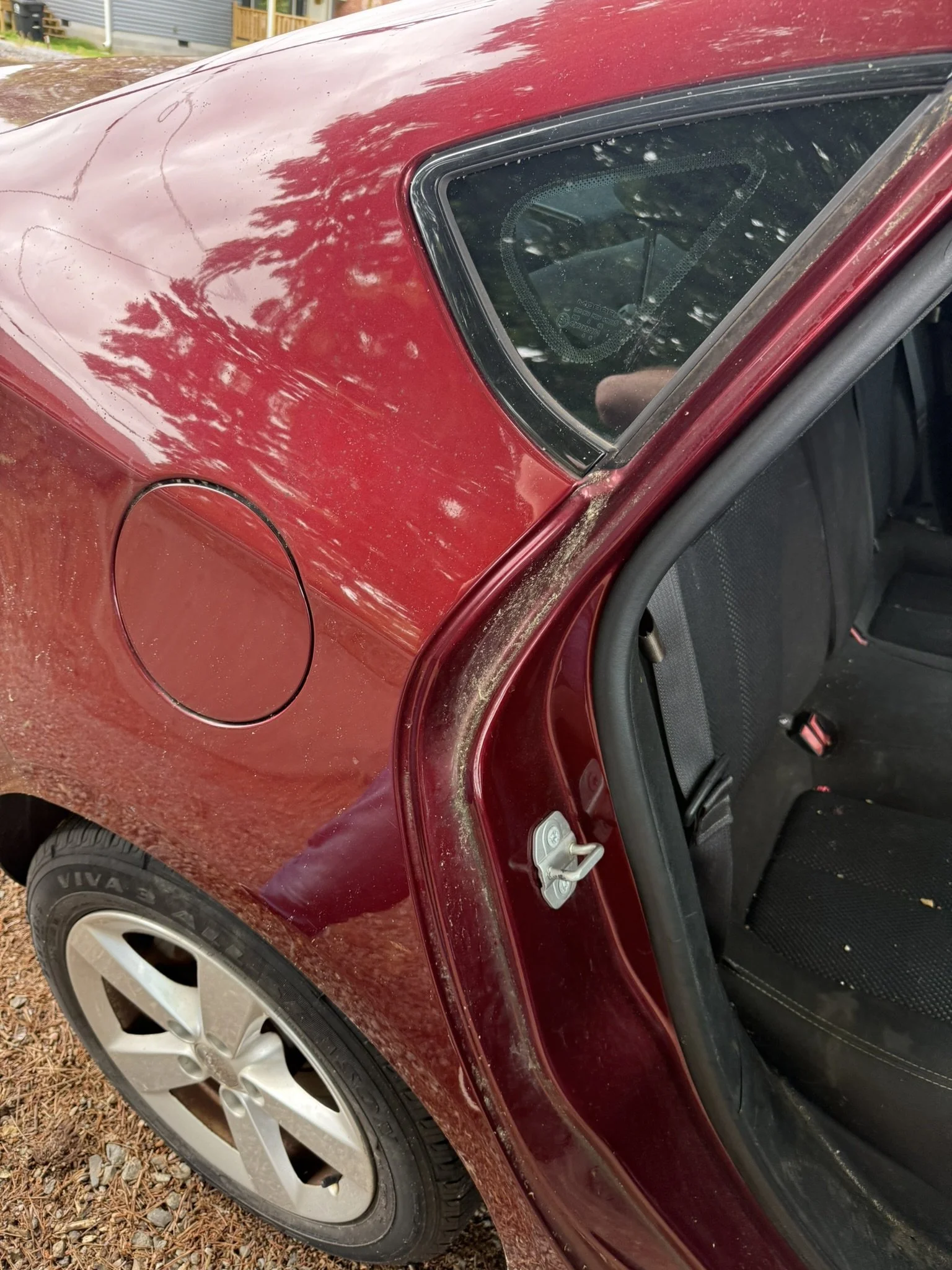 Red car with dirt and mud splatters on the rear quarter panel, tire, and inside the open door, parked on gravel. The rear window shows some dirt and water spots.