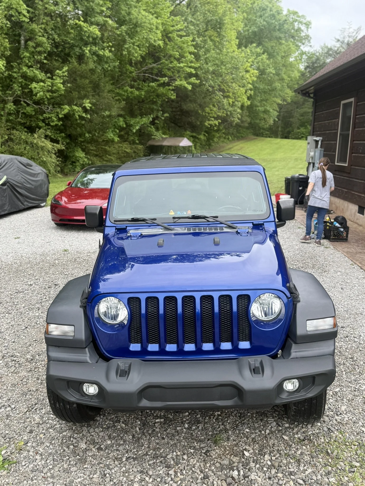 Front view of a blue Jeep parked on a gravel driveway in front of a brown house. A person wearing a gray shirt and jeans is standing near some trash bins on the side of the house. Two other cars, one black and one red, are parked nearby, with a woode