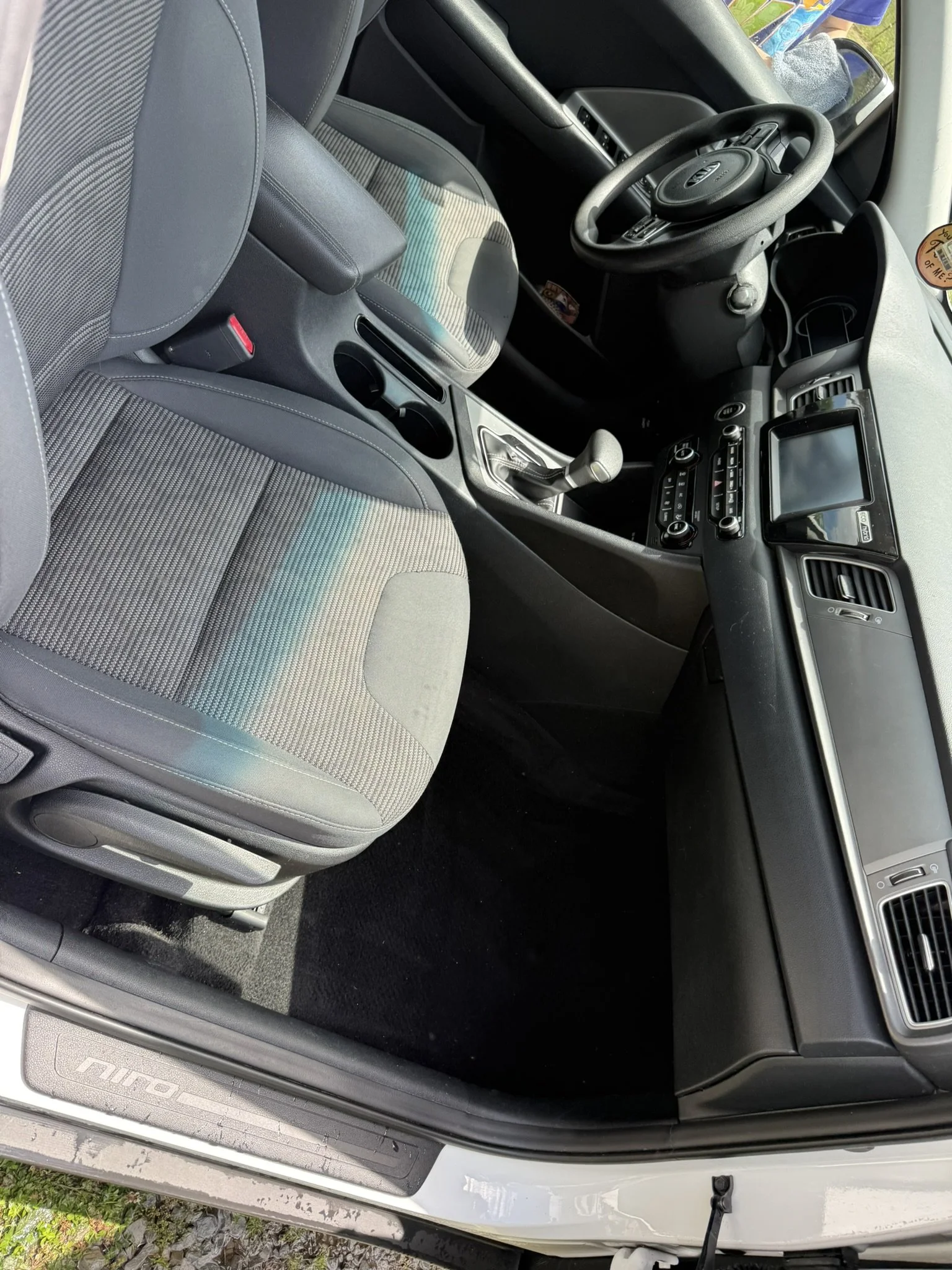 The inside view of a vehicle showing the driver seat, steering wheel, center console with gear shift, and part of the dashboard. There is a person visible through the windshield outside the vehicle.