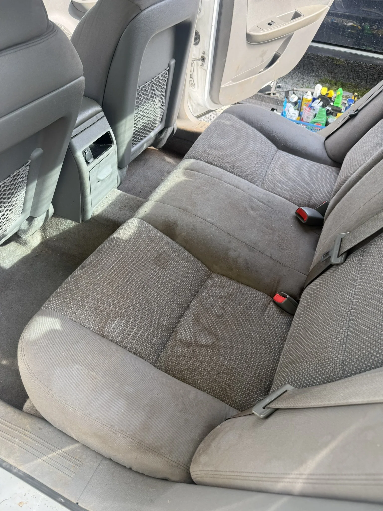 The back seat of a car showing cloth upholstery, seat belts, and the back of the front seats, with a view outside showing cleaning supplies in a container.