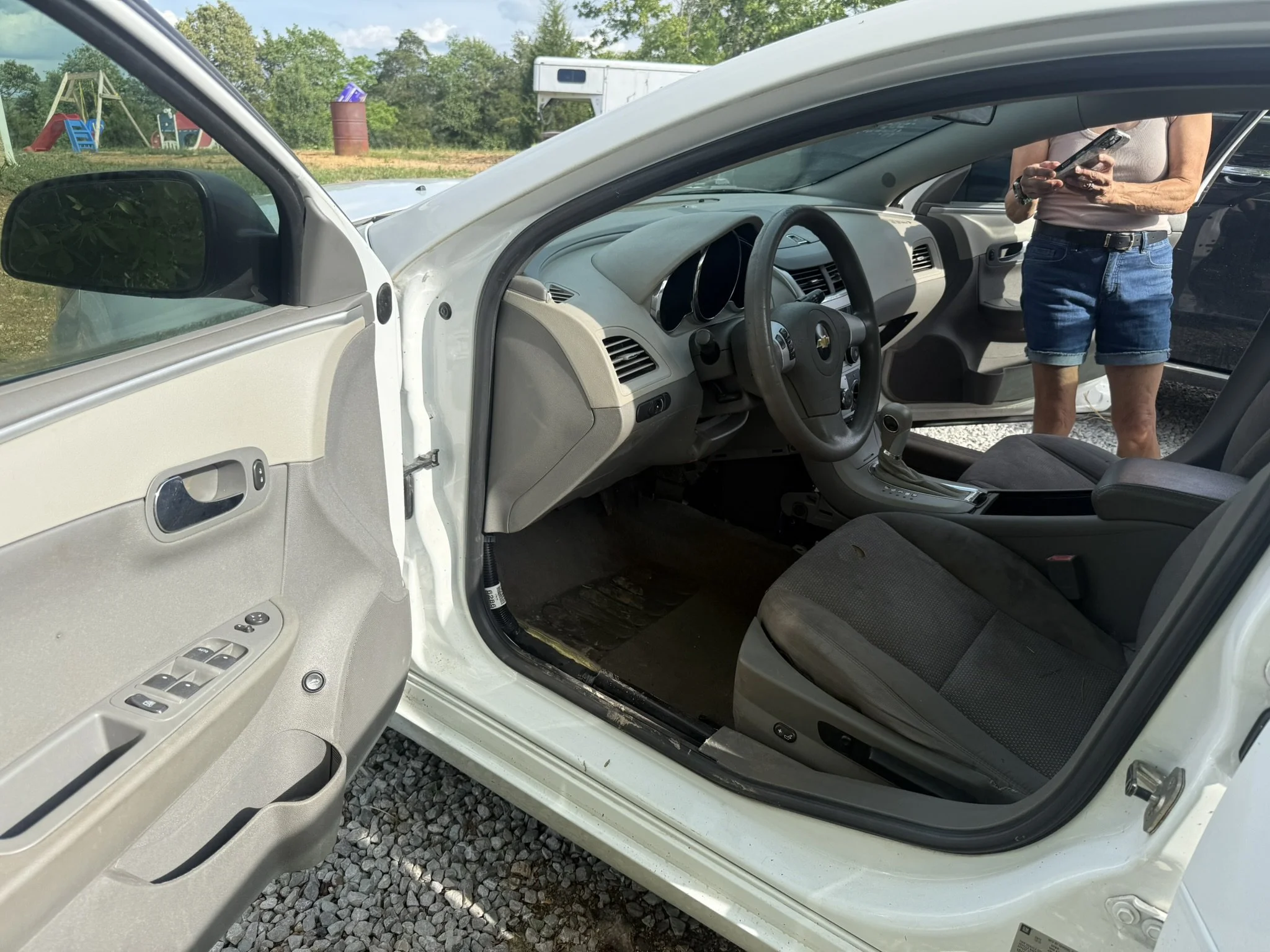 View of the interior of a white car from the driver side, with the door open. A person is standing outside, wearing a sleeveless top and denim shorts, looking and using a smartphone. The background shows a grassy area with trees, a playground with sw