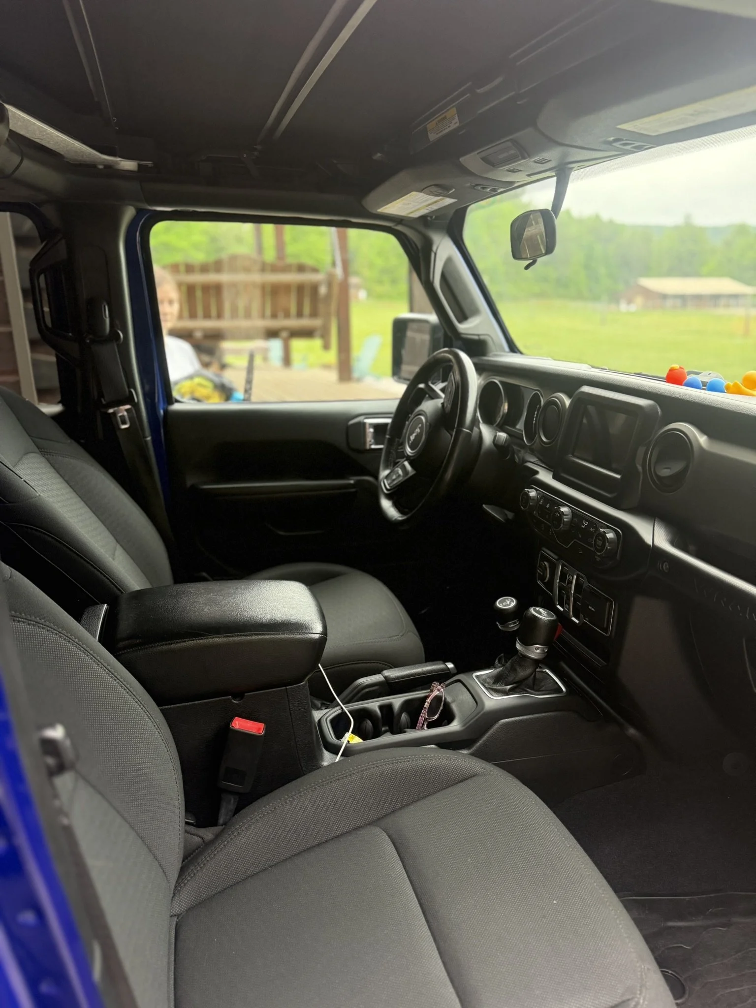 Photo of the interior of a vehicle, showing the front seats, dashboard, steering wheel, gear shift, and a small display screen. The background outside the vehicle shows green grass, trees, and a house.