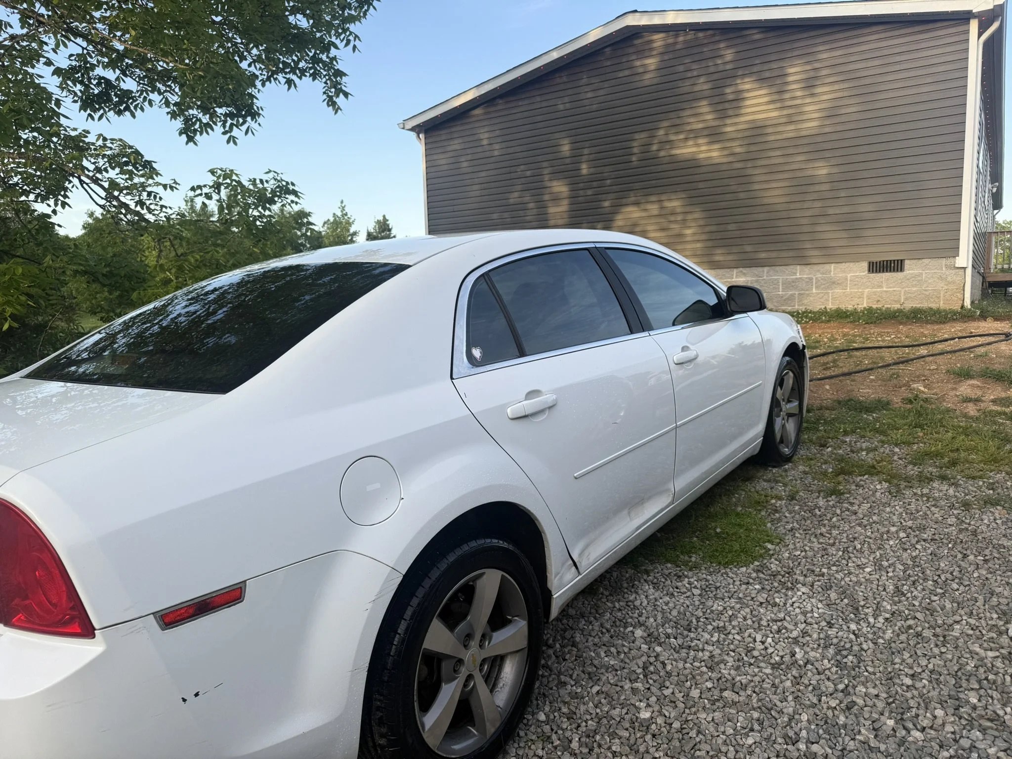 A white car parked on a gravel driveway in front of a brown wooden house with a stone foundation, with trees and a clear blue sky in the background.