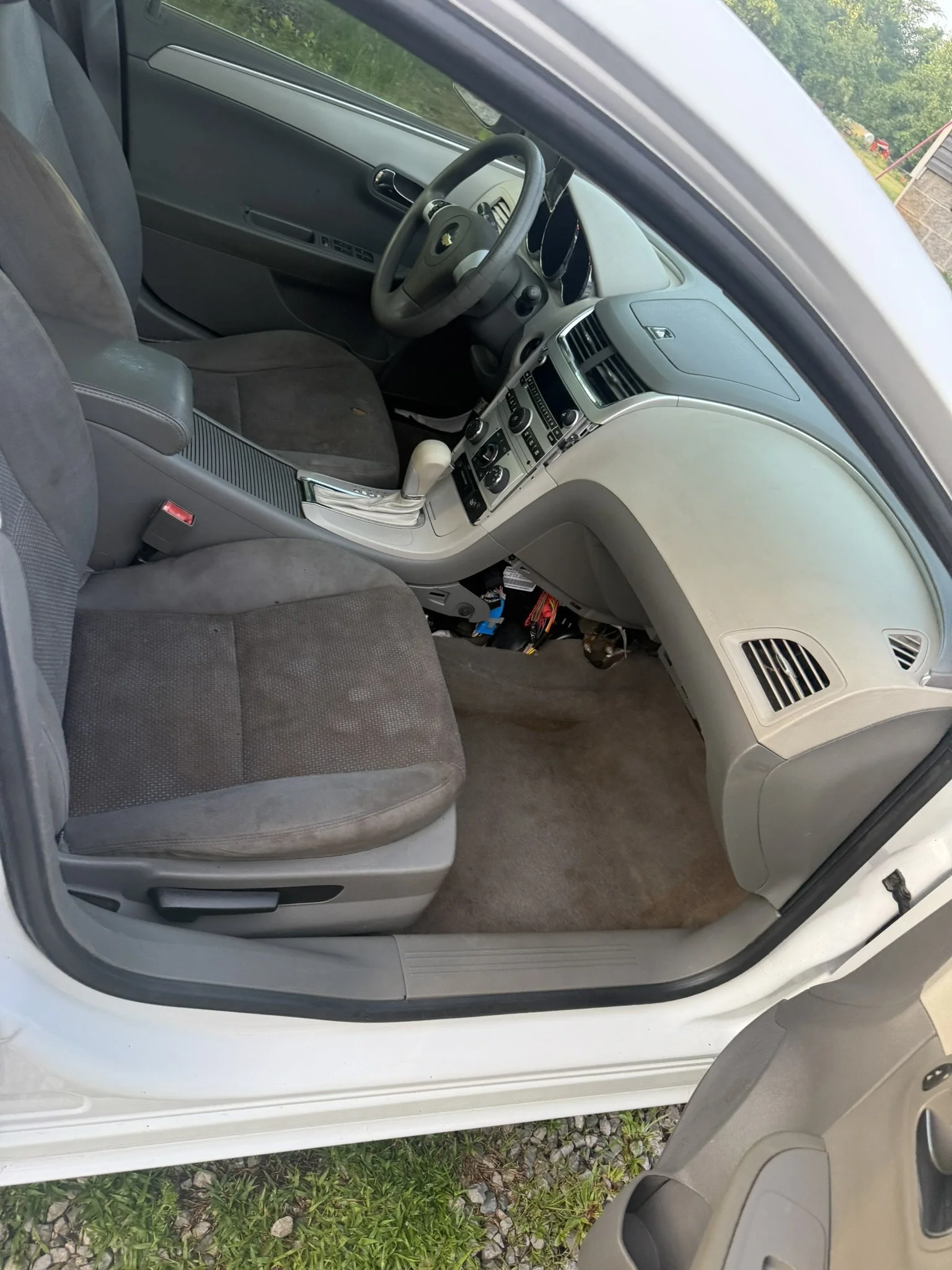 The interior of a Chevrolet car showing the front passenger seat, dashboard, steering wheel, and center console, with green foliage outside the window.