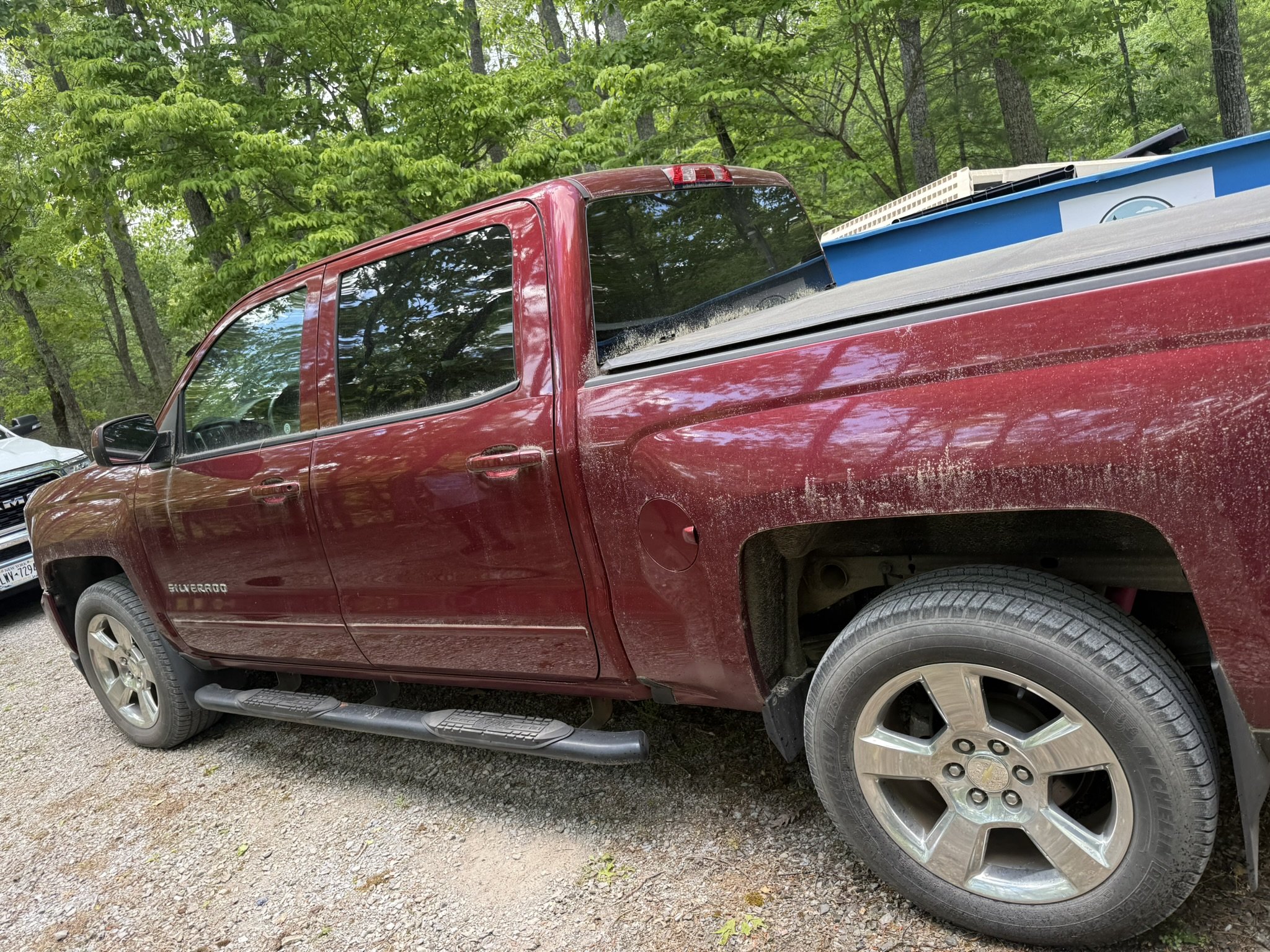 Red pickup truck with loaded cargo bed containing construction or household items, parked on a gravel surface in a wooded area.