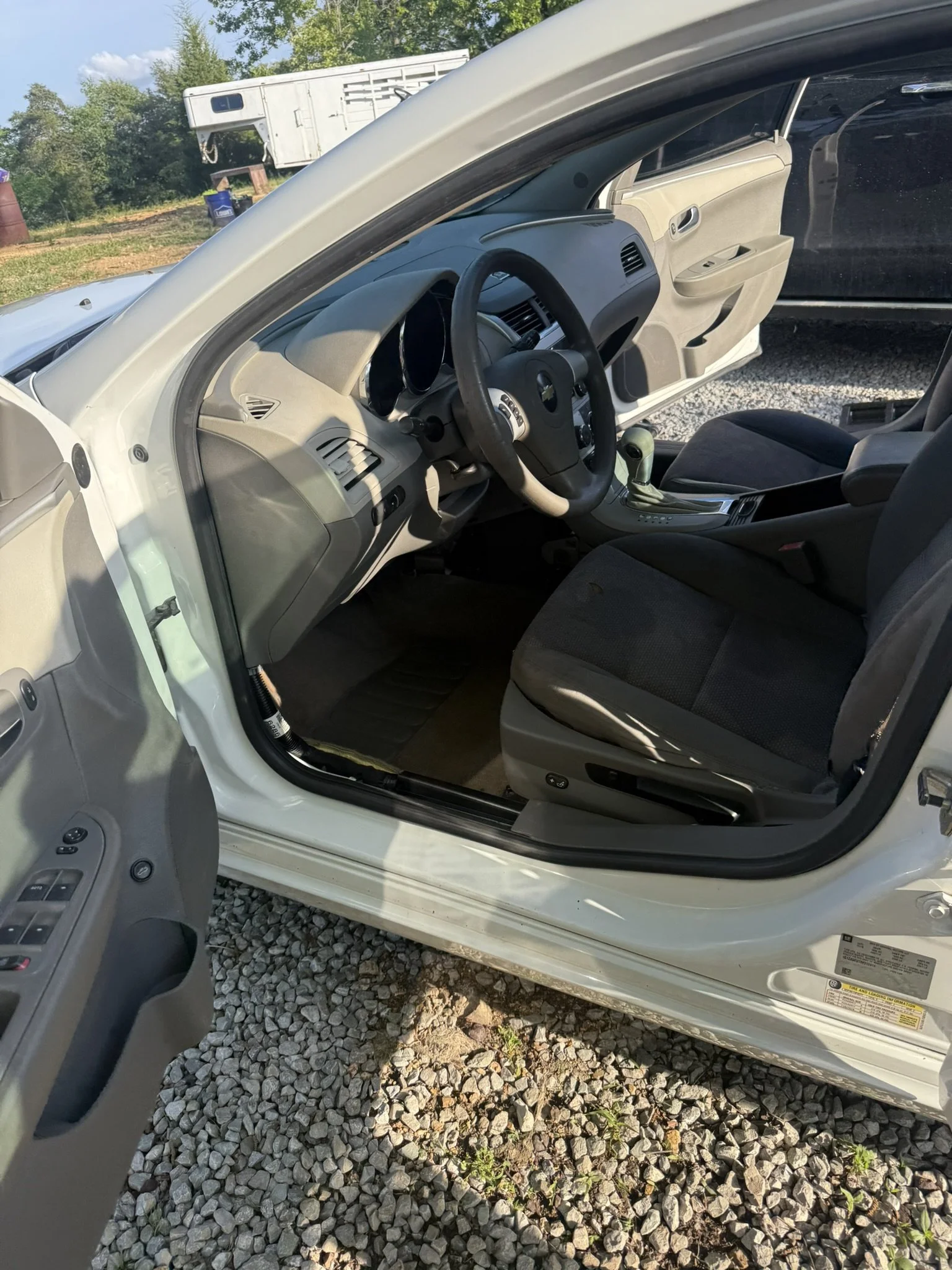 Inside view of a beige car parked on gravel, showing the steering wheel, dashboard, front passenger seat, and open driver's side door with controls.
