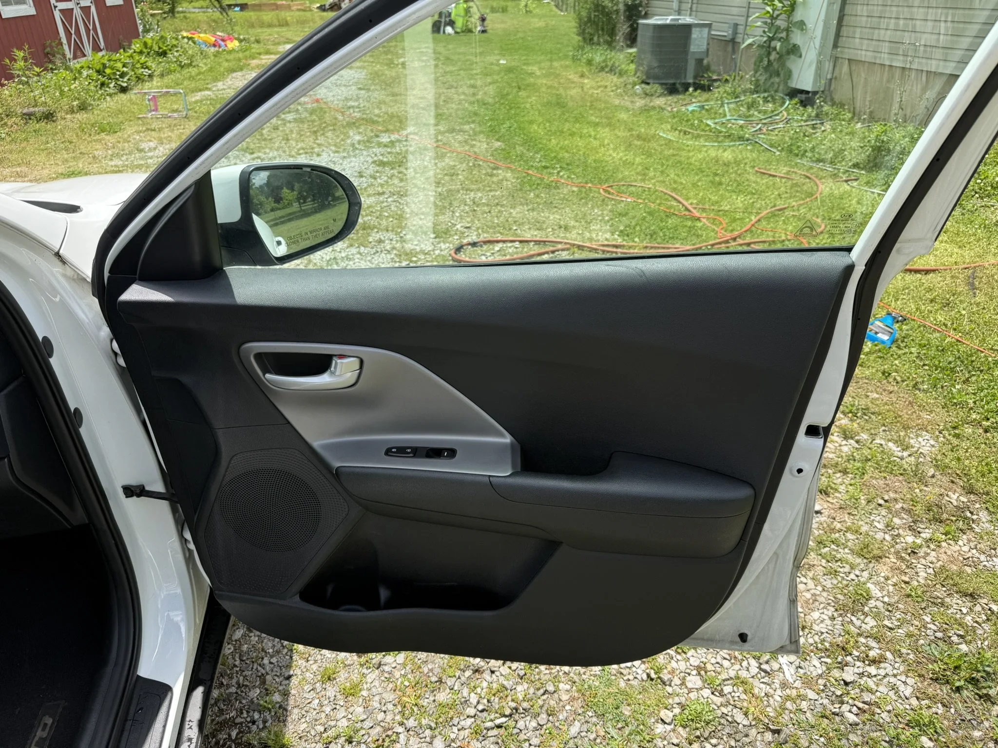 View from inside a vehicle showing the open passenger side door, looking out onto a grassy backyard with a fence, garden, and scattered tools and equipment.
