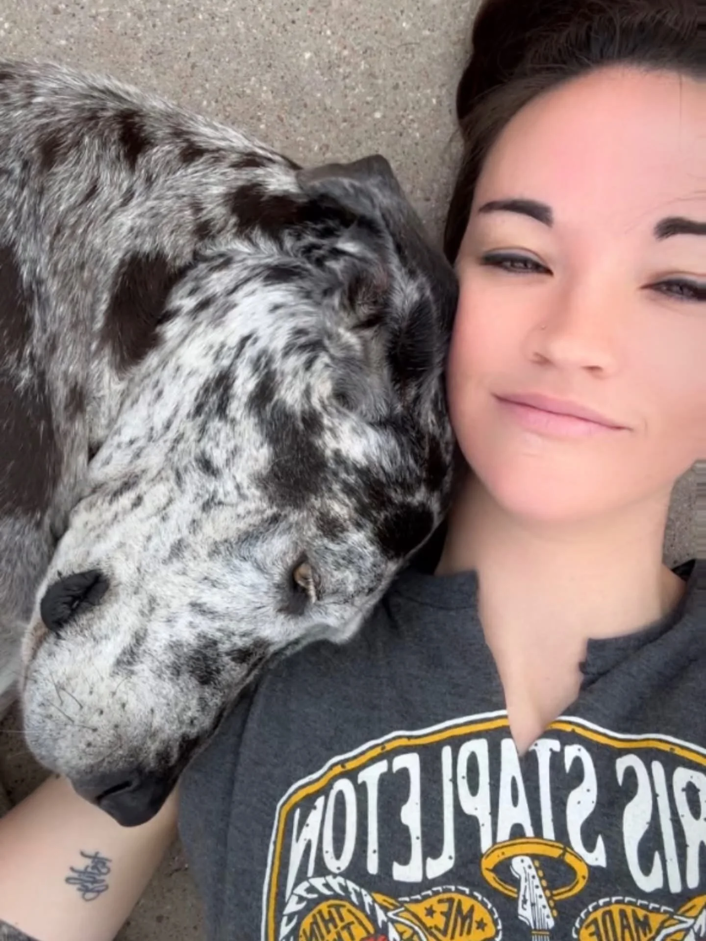 A woman lies on the ground next to a black and white speckled dog, resting her head against the dog. The woman has dark hair and is smiling slightly, wearing a dark shirt with a logo. The ground appears to be dirt or gravel.