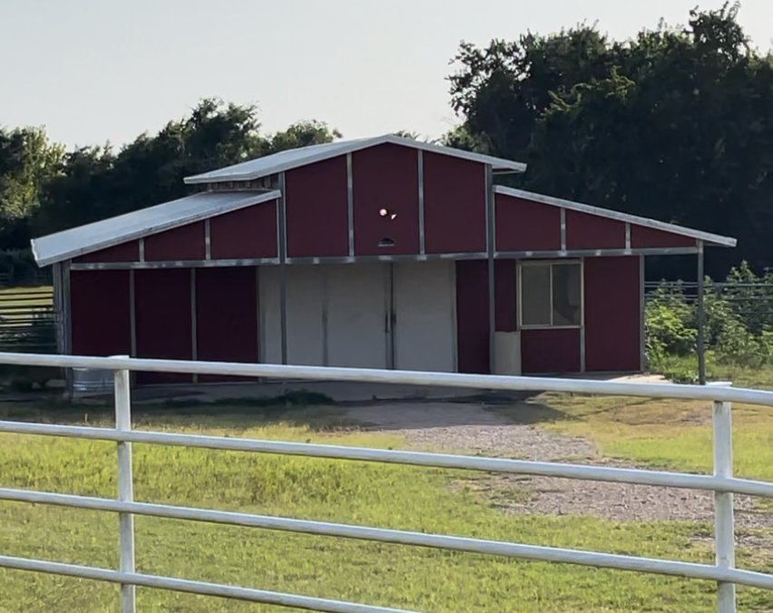 A red barn with silver roof panels and double doors, located on a grassy area with a white fence in the foreground and trees in the background.