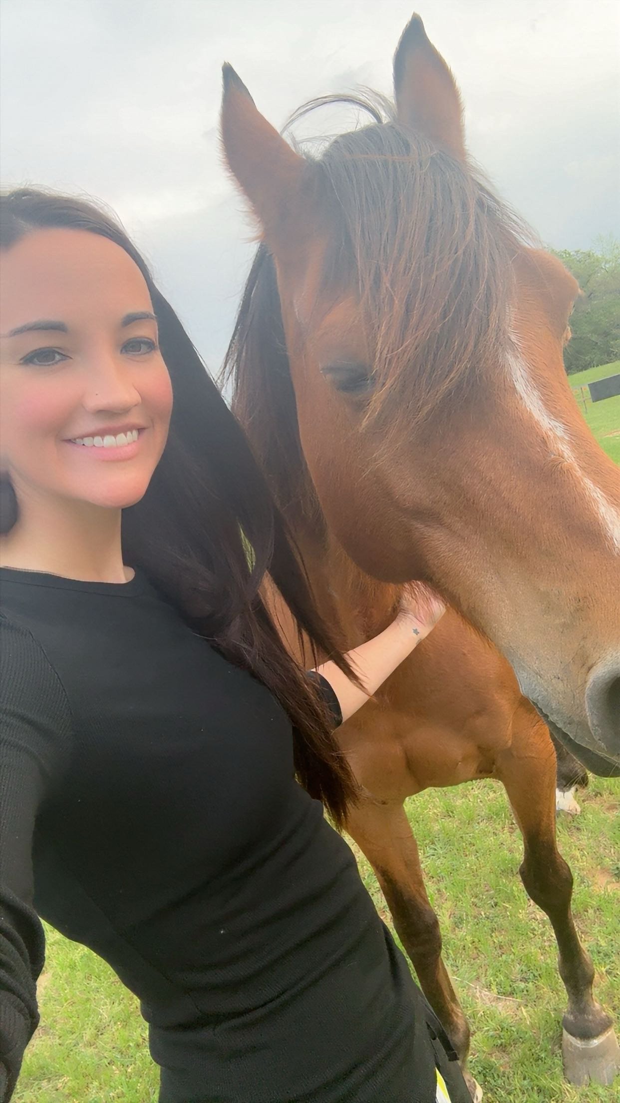 A woman takes a selfie with a brown horse in an outdoor field with grass and trees, overcast sky.
