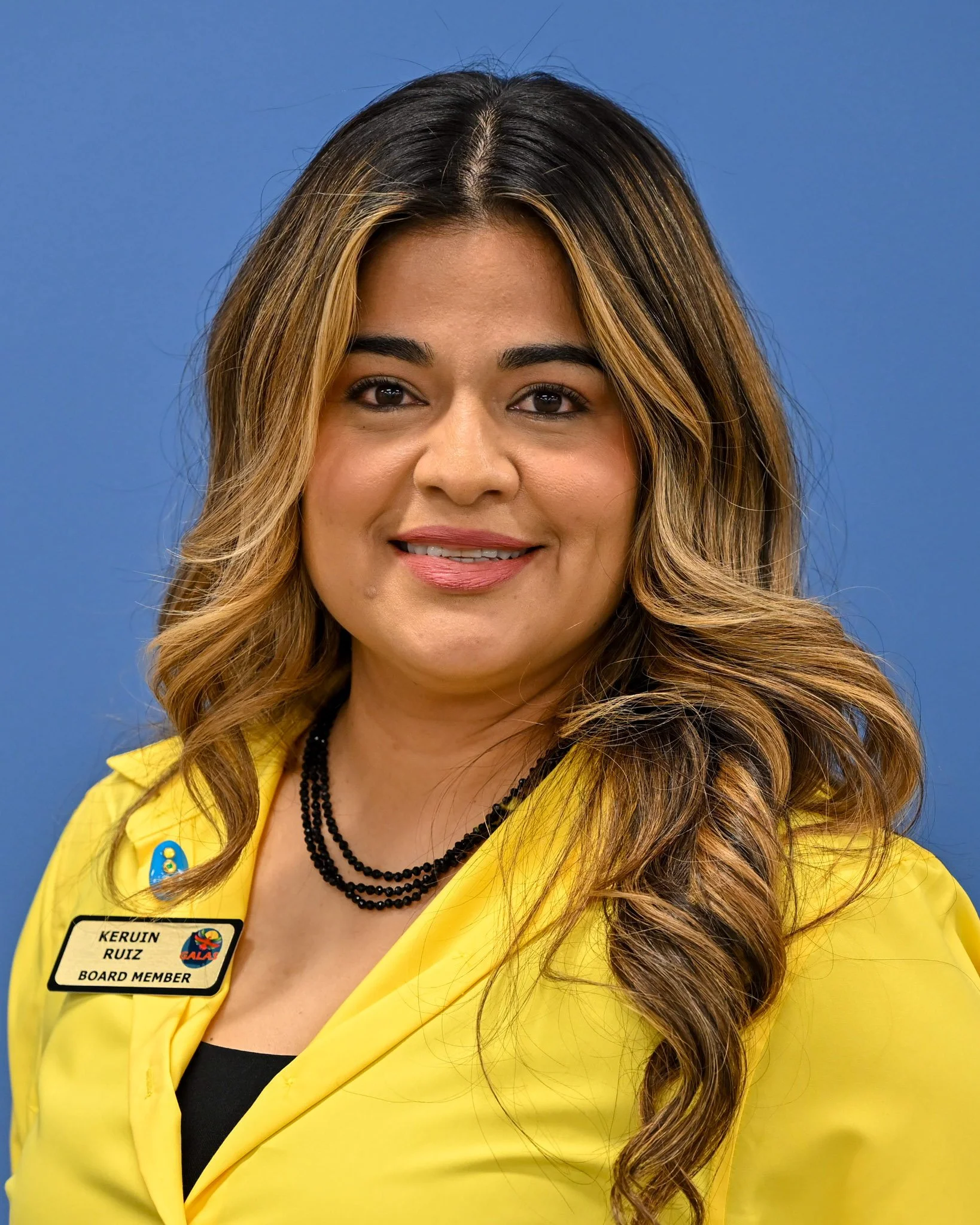 Close-up of a woman with long wavy brown hair with blonde highlights, wearing a yellow blazer, black top, and a name badge that reads 'Kerlin Ruiz Ruiz, Board Member,' and a black beaded necklace, against a blue background.