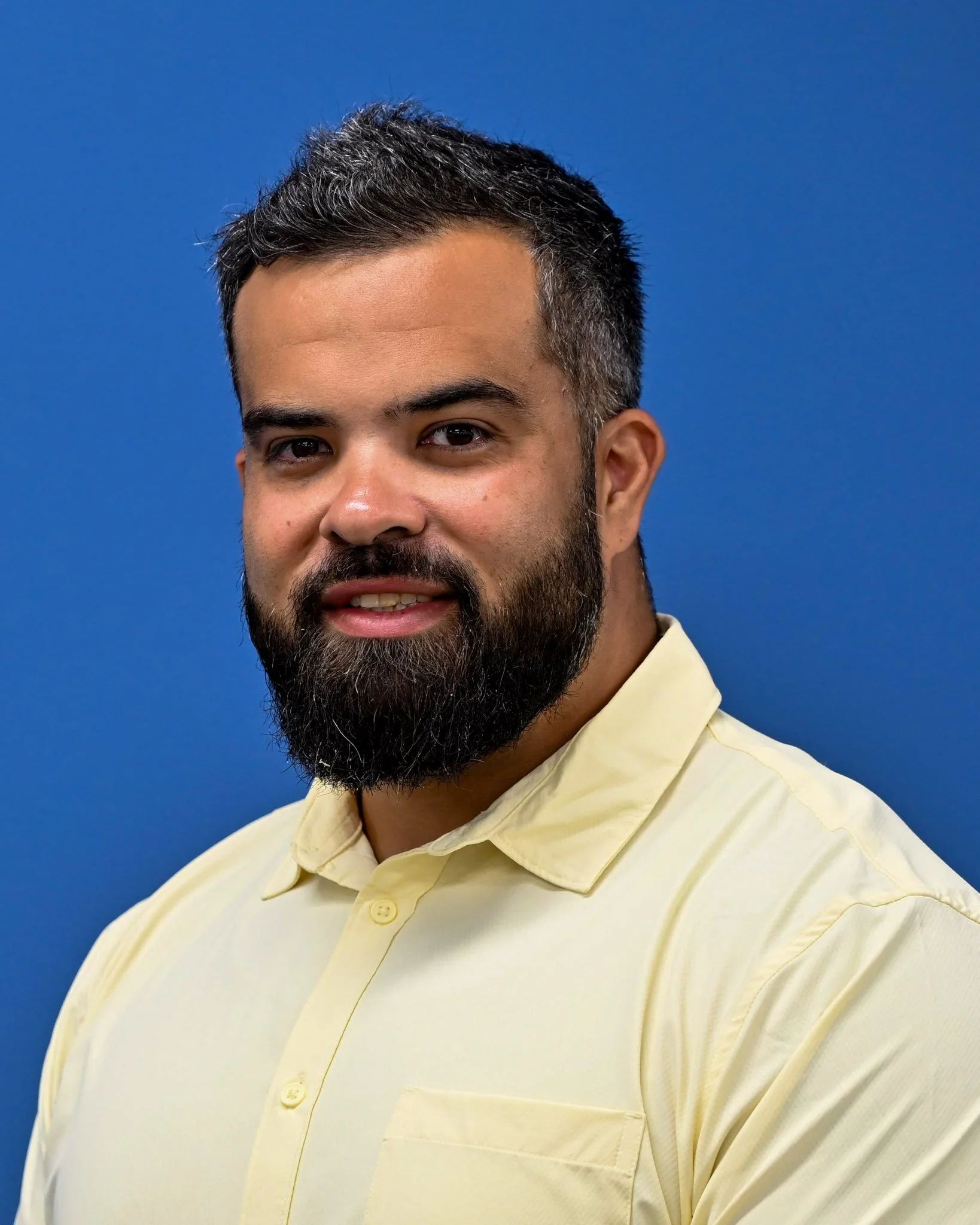 Close-up of a man with dark hair, a beard, and mustache, wearing a yellow collared shirt, with a blue background.