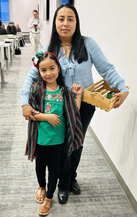 A woman and a young girl standing in a room, with the woman holding a small wooden basket. The girl is smiling and wearing a green top and a striped shawl, while the woman is dressed in a light blue sweater and black pants.