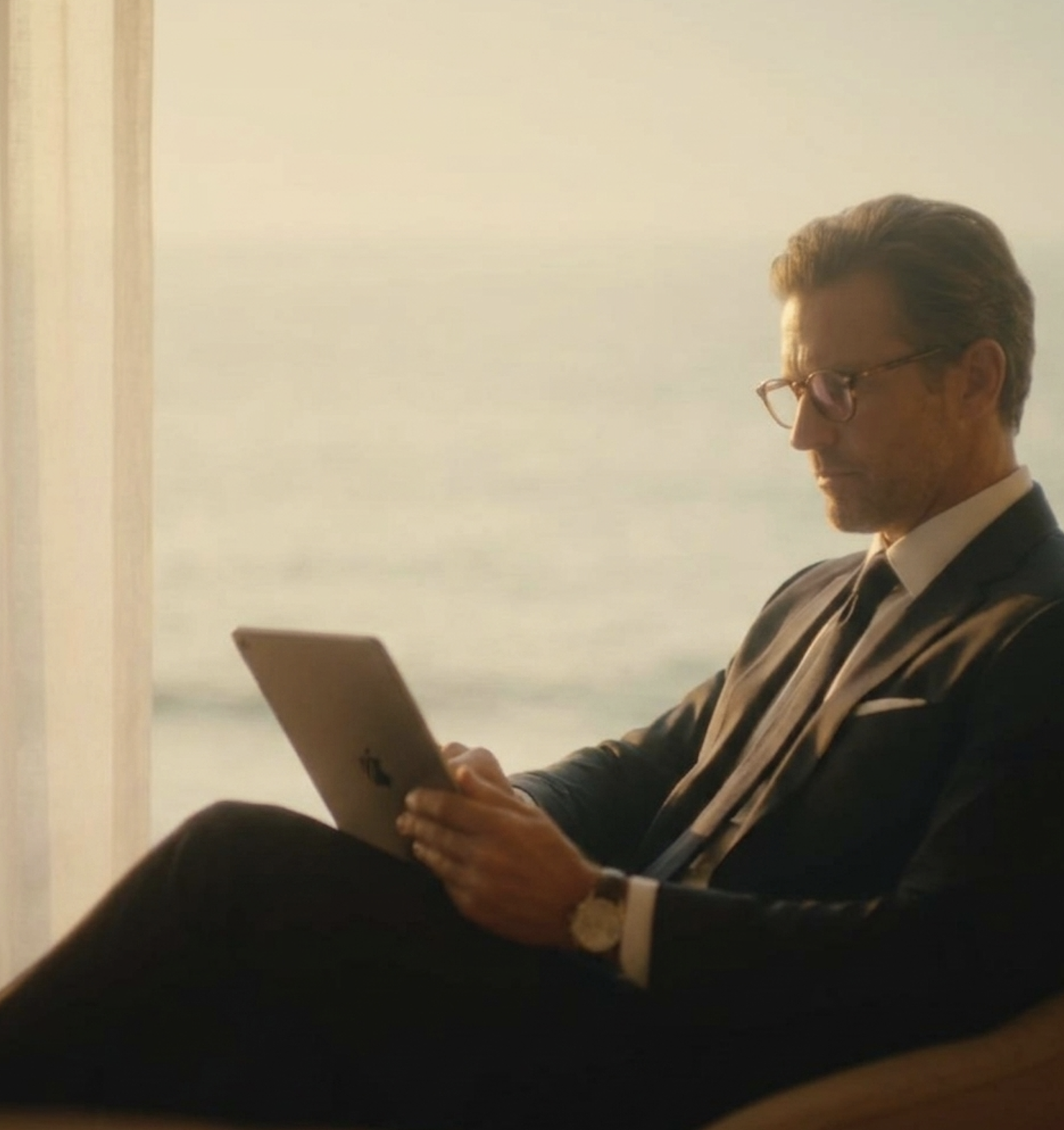 A man with glasses and gray hair sitting indoors, looking at a tablet device.