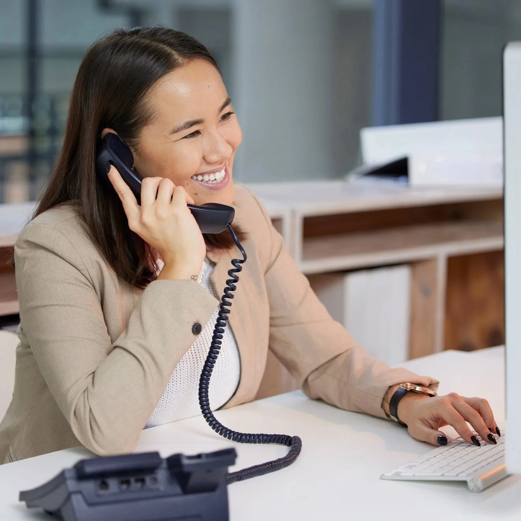 A woman with dark brown hair, wearing a beige blazer and a white top, smiling while talking on a landline phone in an office environment. She is also using a computer keyboard with her right hand, and a smart watch is visible on her left wrist.