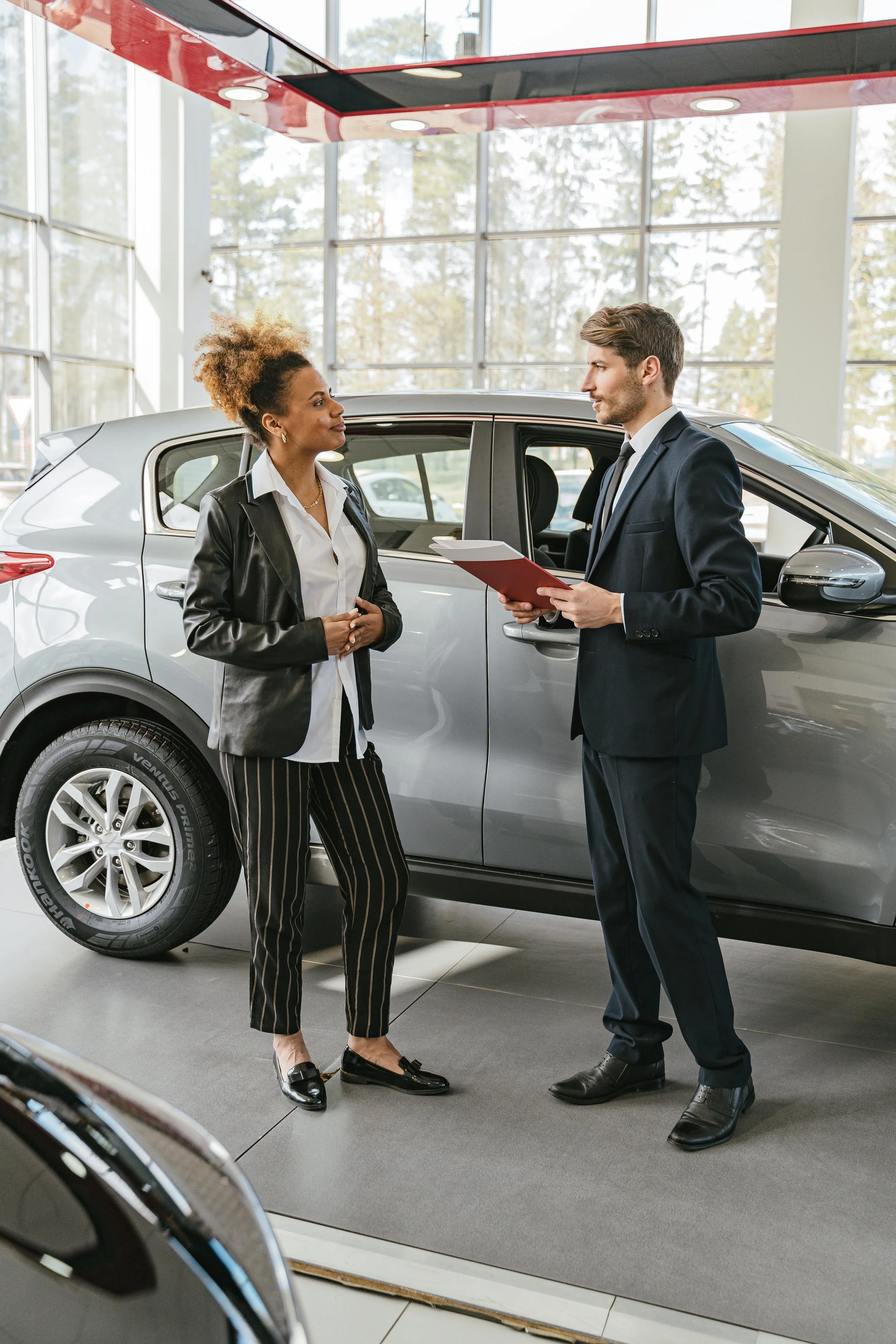 A woman and a man inside a car dealership having a conversation. The woman is wearing a black leather jacket, white shirt, and striped pants, while the man is wearing a dark suit and holding a folder. There are cars and large windows in the background.