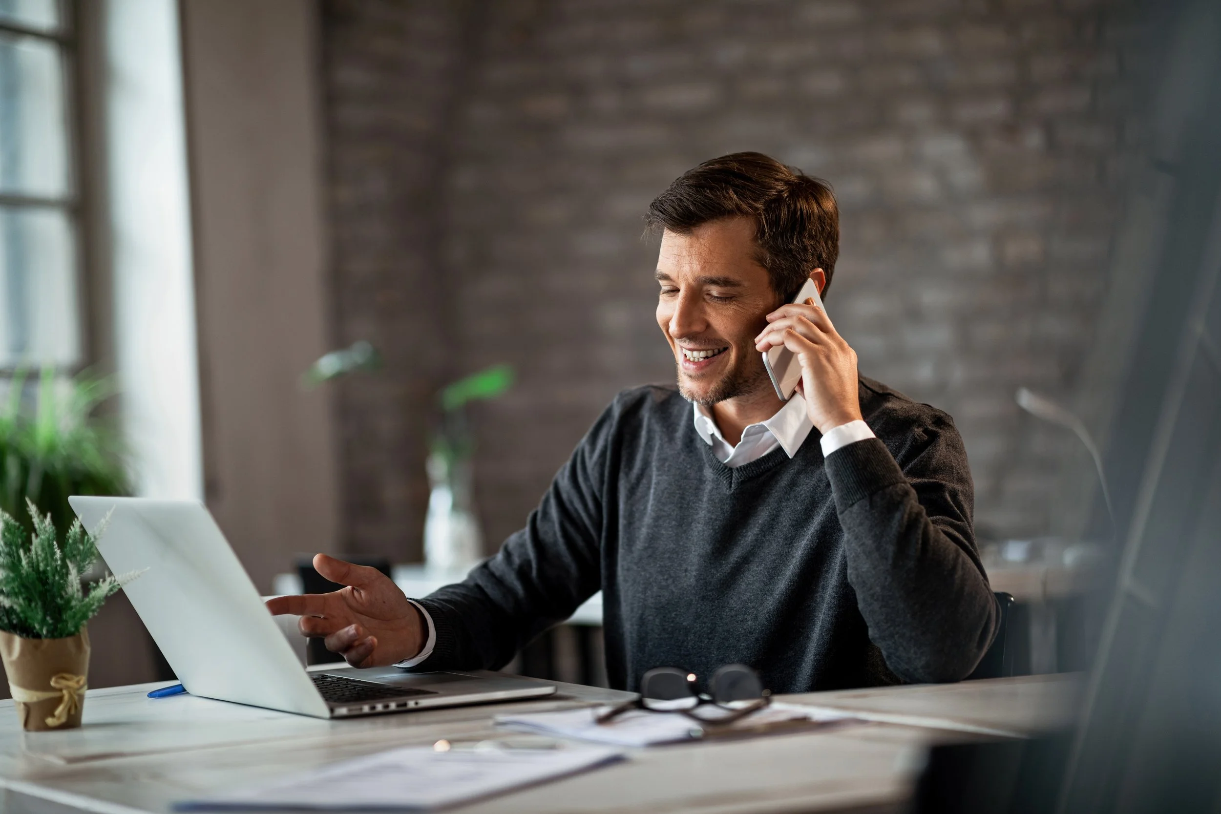 A man with dark hair and a beard, wearing a black sweater over a white shirt, smiling while talking on a cellphone at a desk with a laptop, glasses, and papers, in a modern office with a brick wall background and natural light.