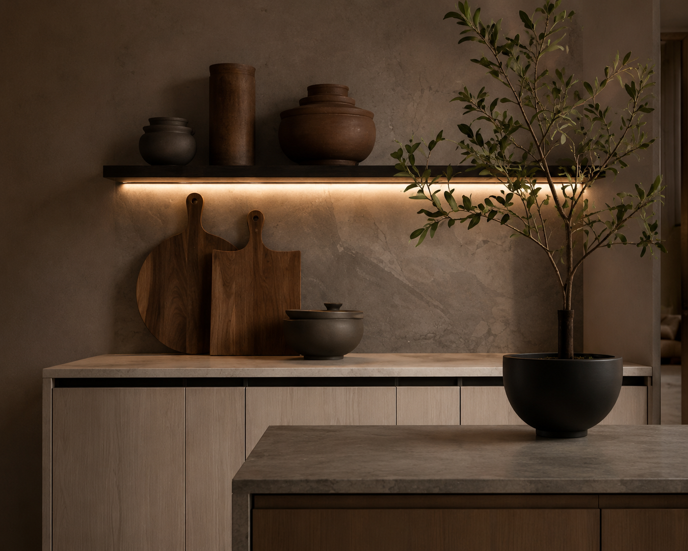 Modern interior kitchen with a minimalist design, featuring a light wood cabinet, a gray countertop, decorative wooden cutting boards, a black bowl with a lid, a potted tree, and warm ambient lighting.