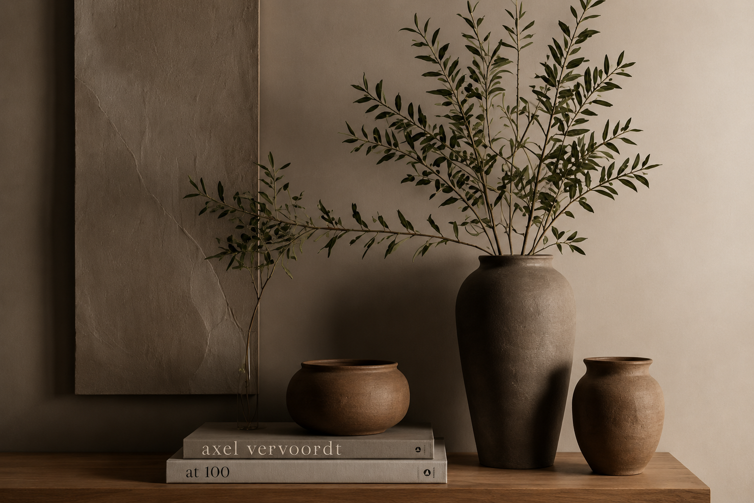 Decorative image with a large brown vase with green leafy branches, a smaller brown vessel, and a book titled 'axel vervoordt at 100' on a wooden surface against a neutral wall with abstract art.