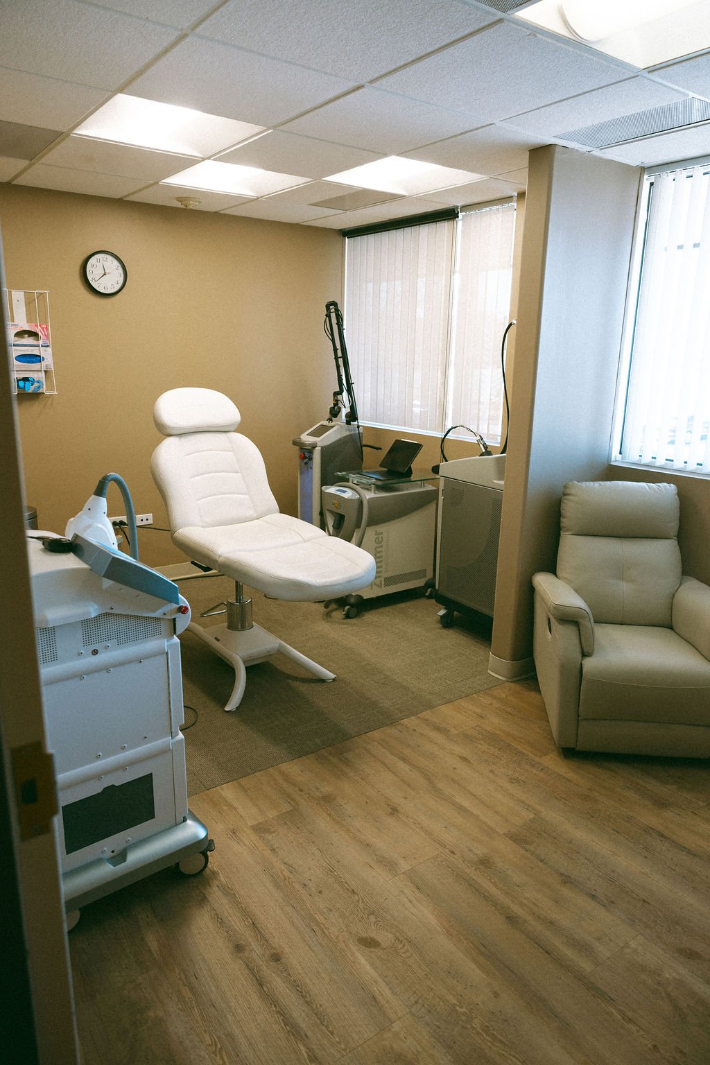 A medical examination room with a white examination chair, medical equipment, a beige wall clock, and a cream-colored armchair near large windows with vertical blinds.